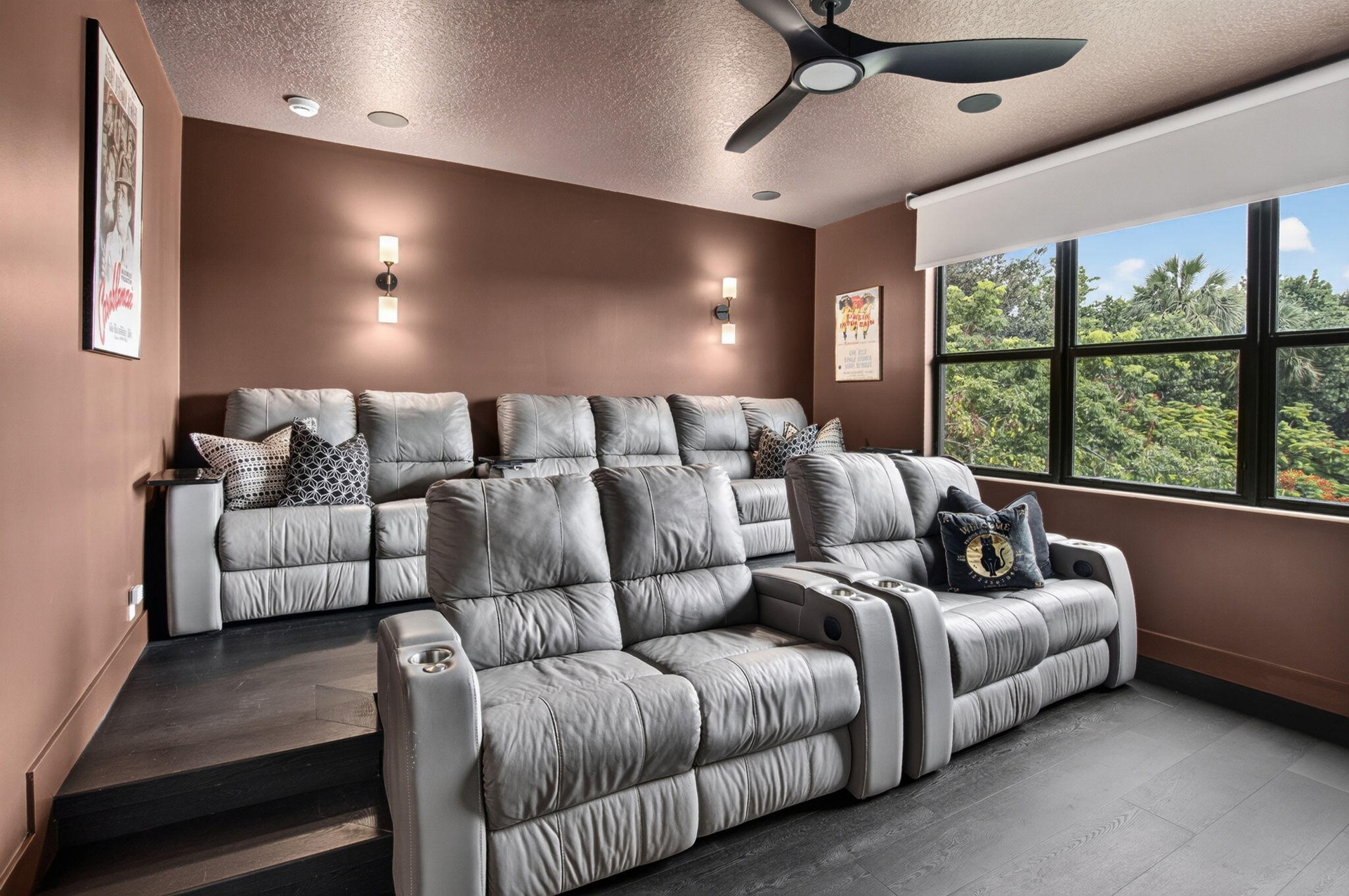 Dedicated media room featuring tiered seating, built-in wall sconces, a modern ceiling fan, and large black-framed windows overlooking mature trees