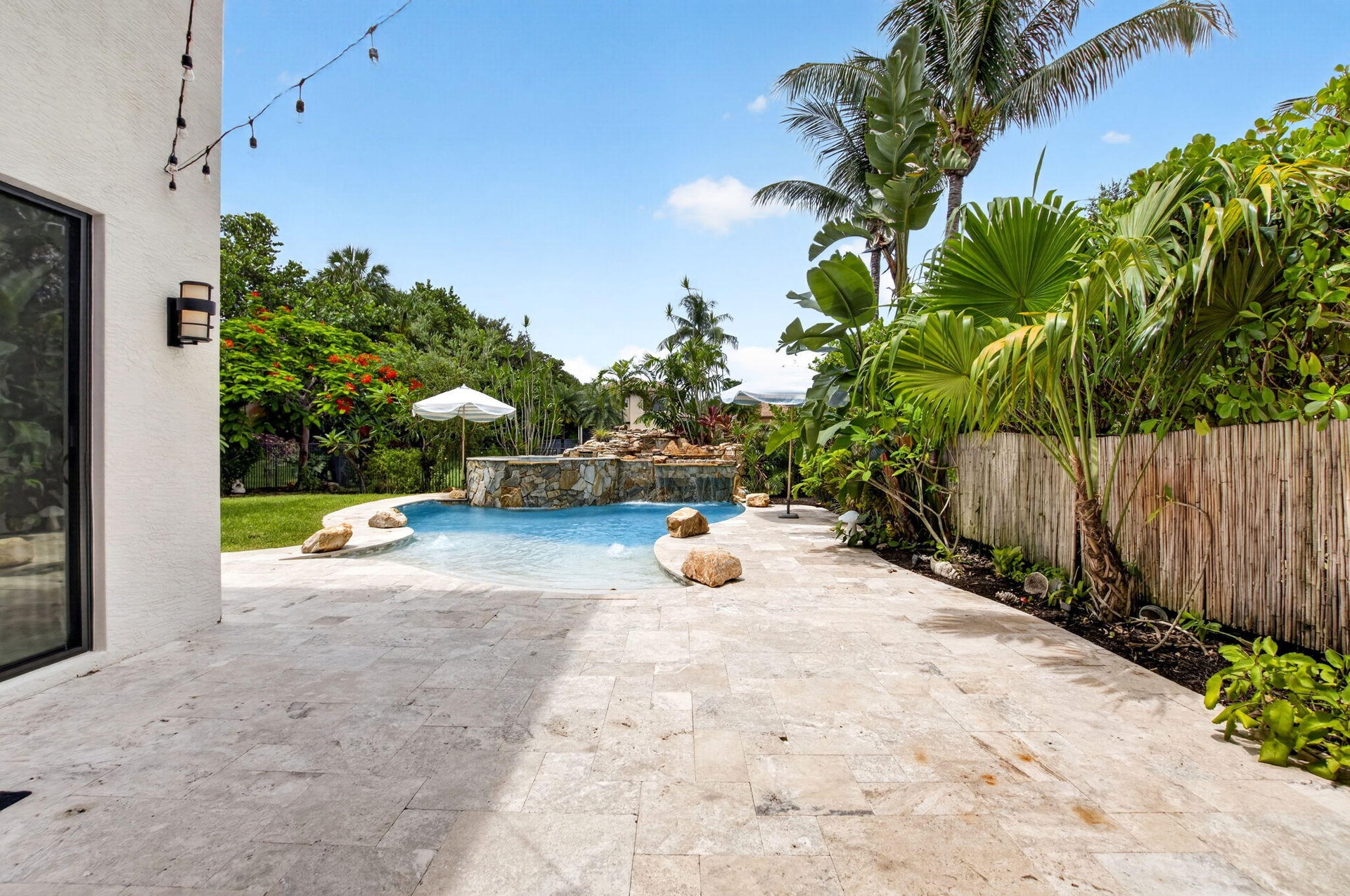 Expansive patio featuring light-toned natural stone pavers, a free-form swimming pool with a rock waterfall feature, tropical landscaping, and a bambo