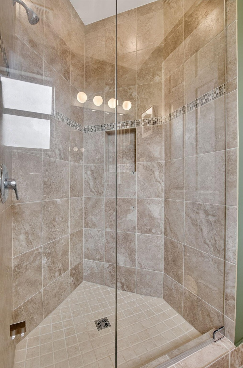 Glass-enclosed shower featuring large format wall tiles, a decorative mosaic accent band, a built-in shower niche, and a corner window