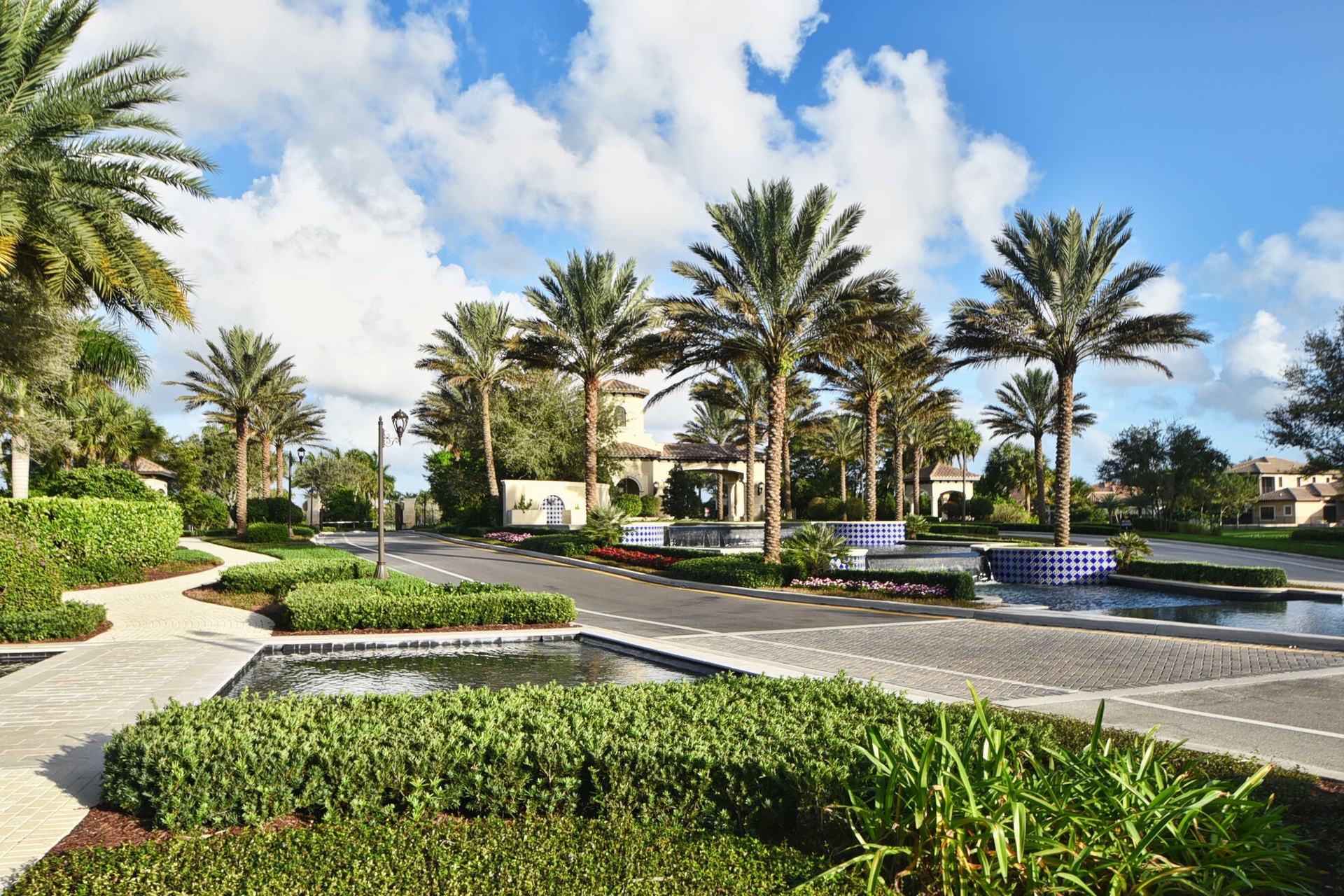 Entryway featuring paver roads, lush landscaping, multiple water features, and mature palm trees