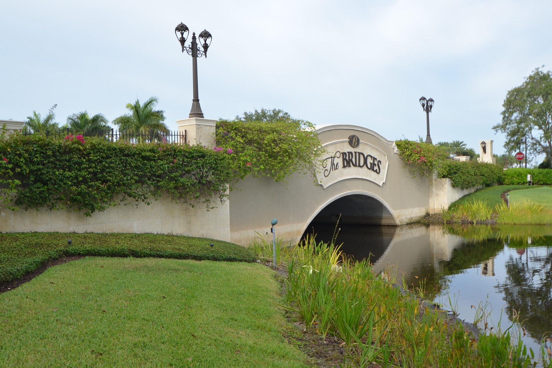 The Bridges entrance bridge featuring decorative street lamps, lush green landscaping, and reflective water