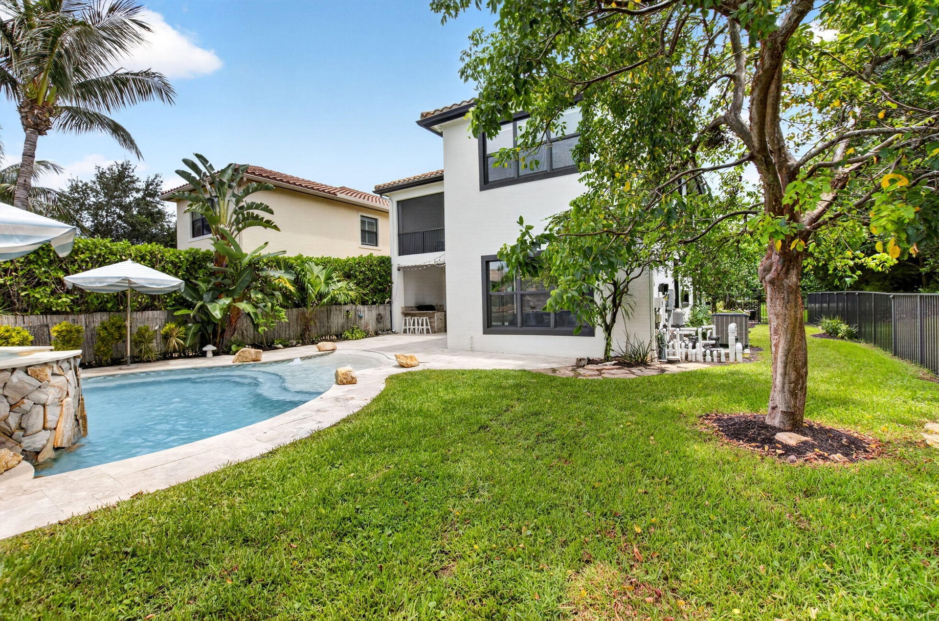 Expansive backyard featuring a curved swimming pool with a stone-clad waterfall, a large grassy area, mature trees, and a covered patio