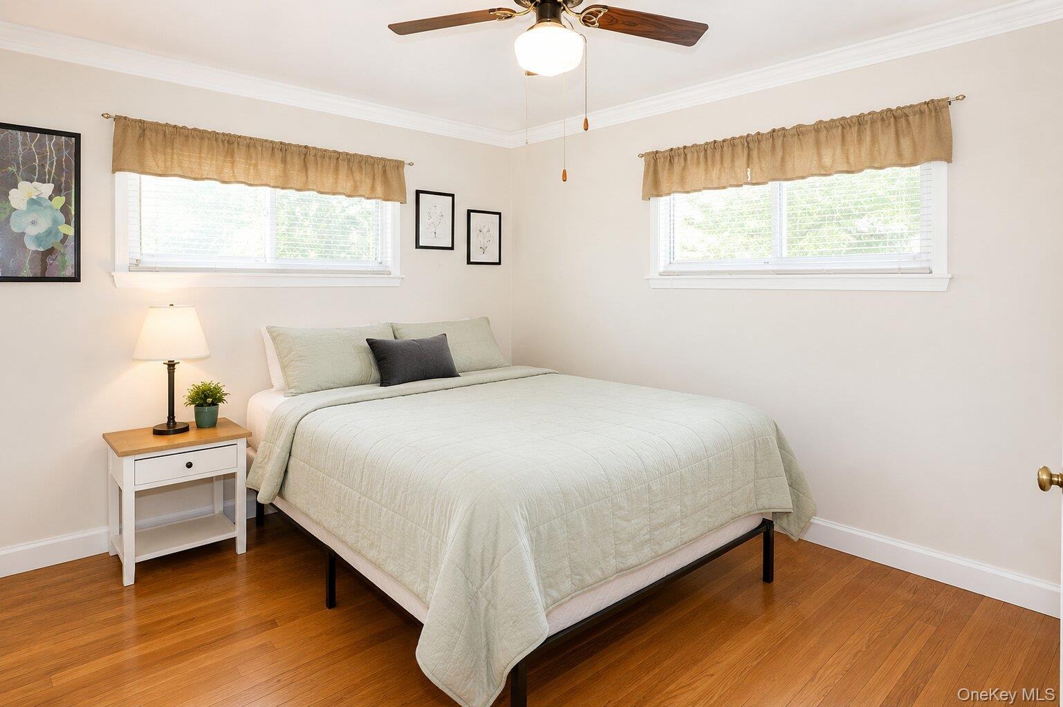 VIRTUALLY STAGED. Bright bedroom featuring wood-finish flooring, two windows, and a ceiling fan with integrated lighting