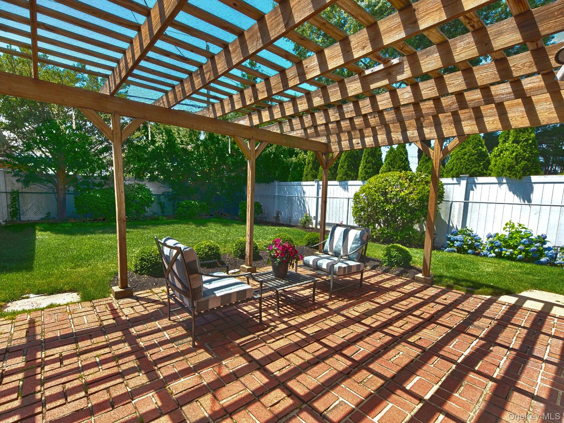 Brick paver patio featuring a wooden pergola with translucent roofing panels, surrounded by a manicured lawn, mature shrubs, and fencing