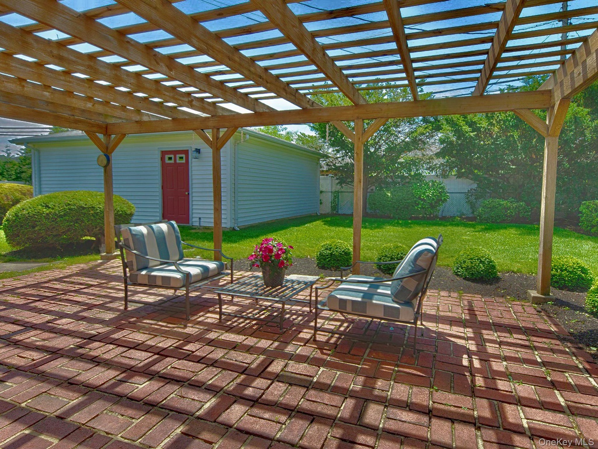 Brick paver patio featuring a wooden pergola with translucent roofing panels, surrounded by a manicured lawn, mature shrubs, and fencing