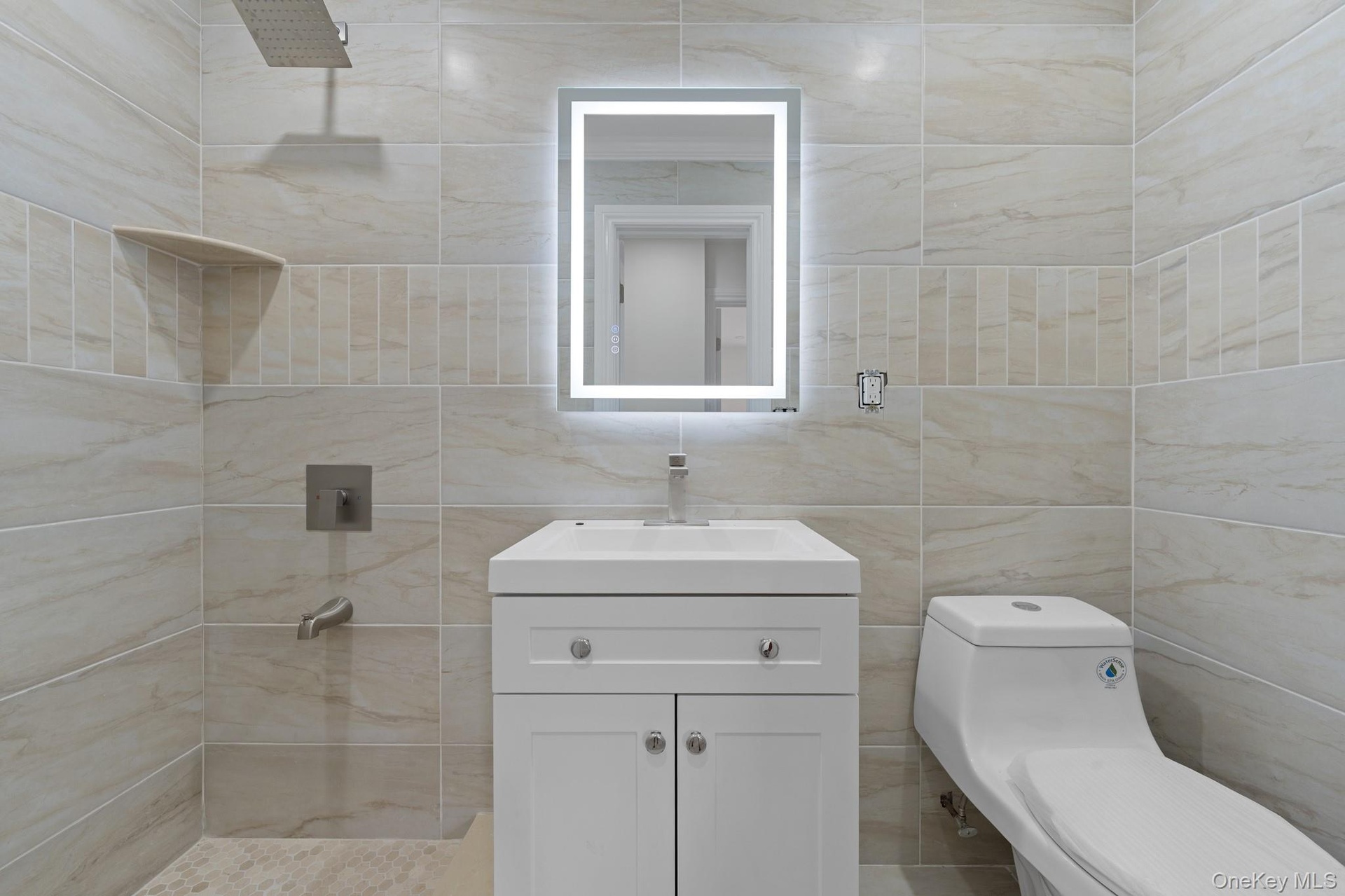 Bathroom featuring large format light-toned tile, a rectangular illuminated mirror, a white vanity with under-sink storage, a wall-mounted shower syst