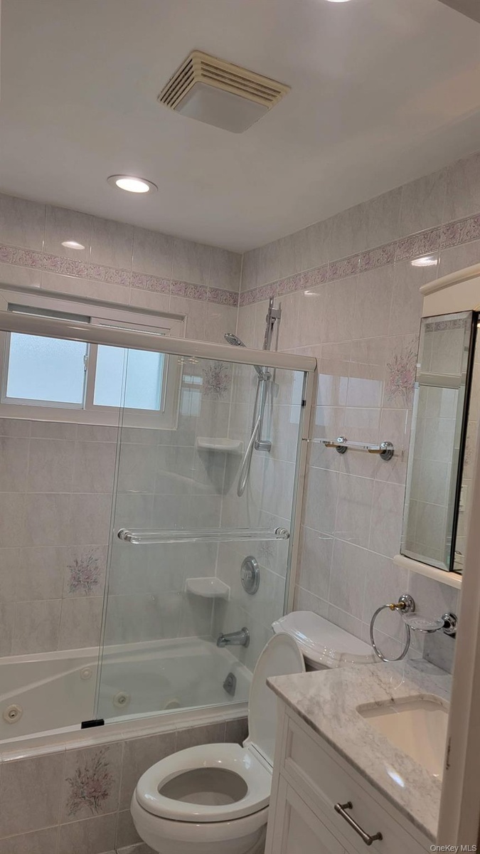 Bathroom featuring a jetted tub with glass enclosure, white vanity with stone countertop, recessed lighting, and tiled walls with decorative accents