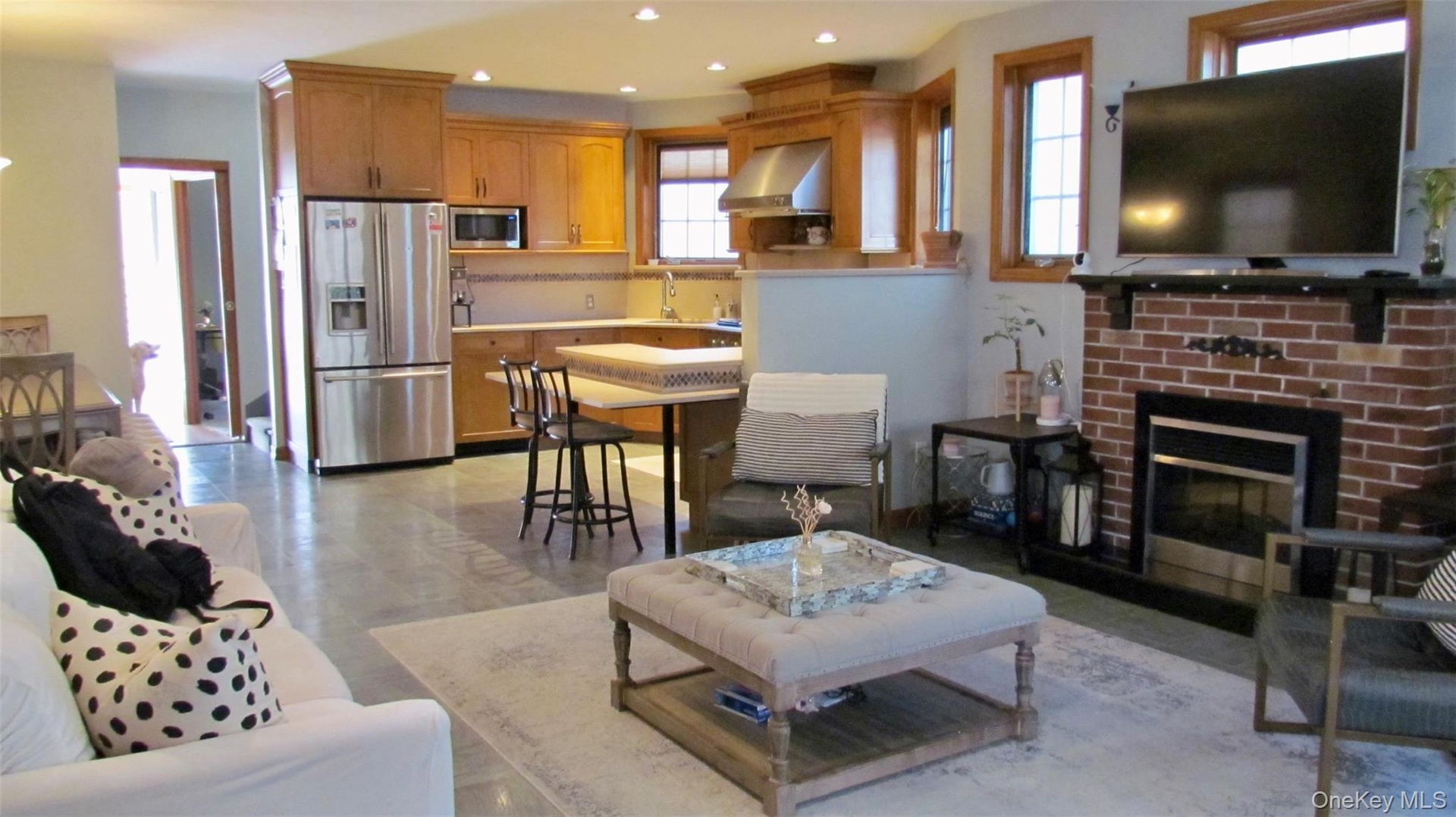 Open-concept living space featuring a brick fireplace with a dark mantel, wood-finish flooring, and natural wood window trim