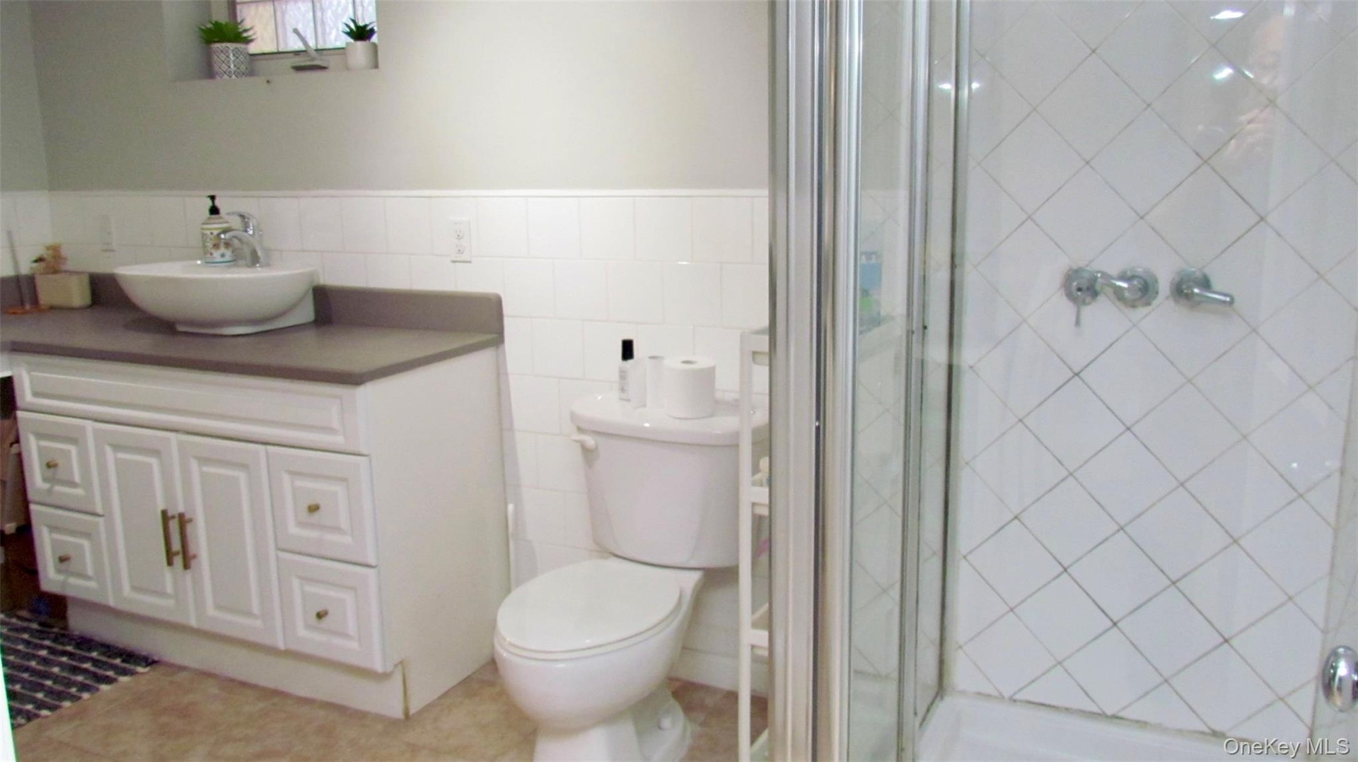 Bathroom featuring a walk-in shower with glass enclosure, white tile walls, a white vanity with vessel sink, and a toilet