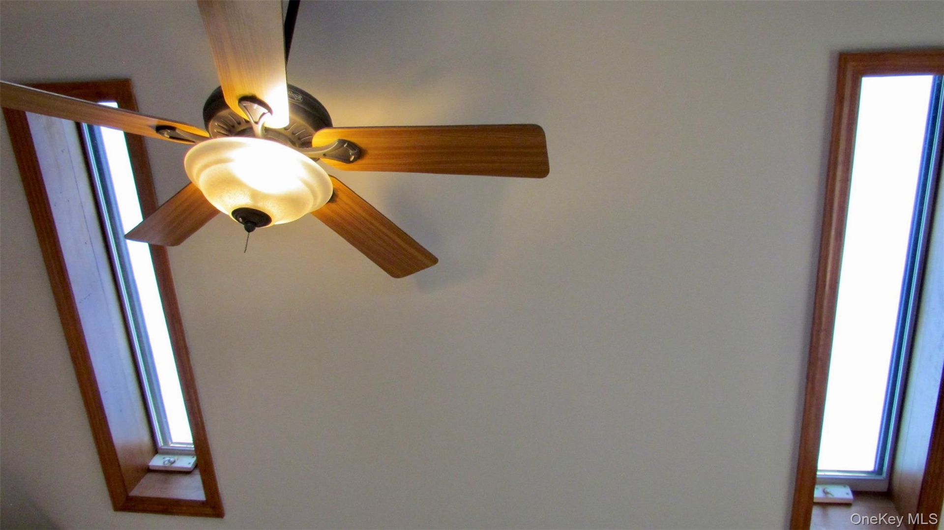 Ceiling fan with light fixture and wood-finish blades, flanked by two elongated skylights with wood trim