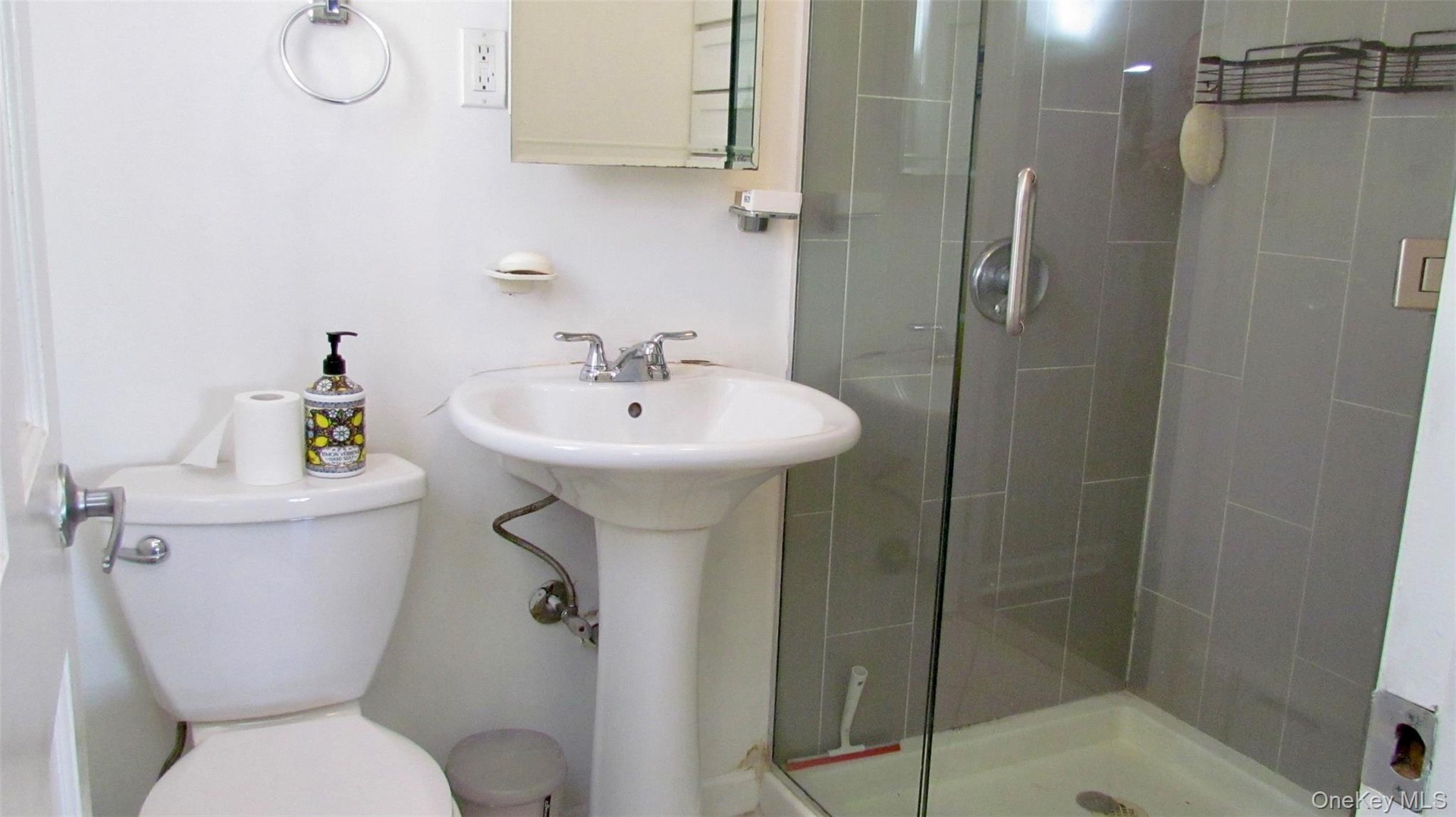 Bathroom featuring a pedestal sink with chrome faucet, toilet, and a glass-enclosed shower with gray tiled walls