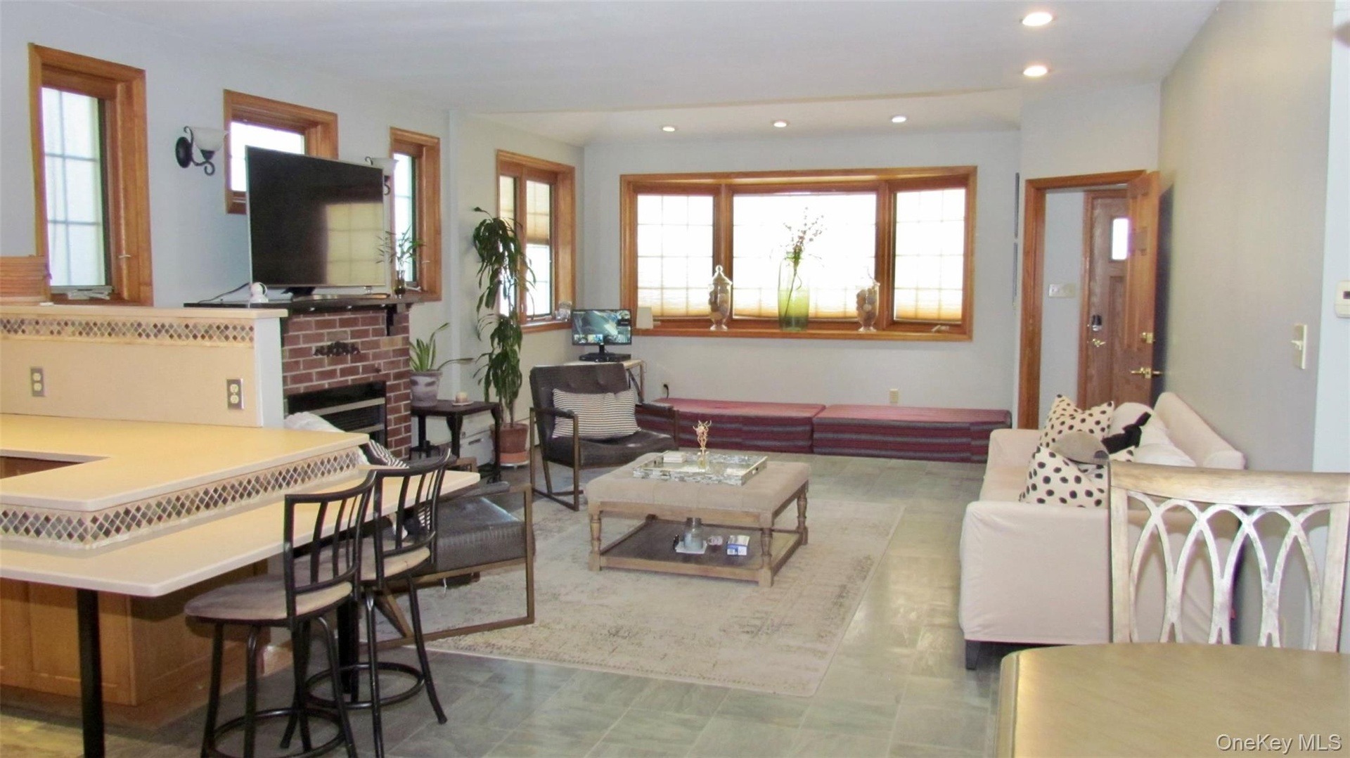 Open concept living area featuring tile flooring, a brick fireplace, built-in breakfast bar with patterned trim, wood-framed windows, and recessed lig