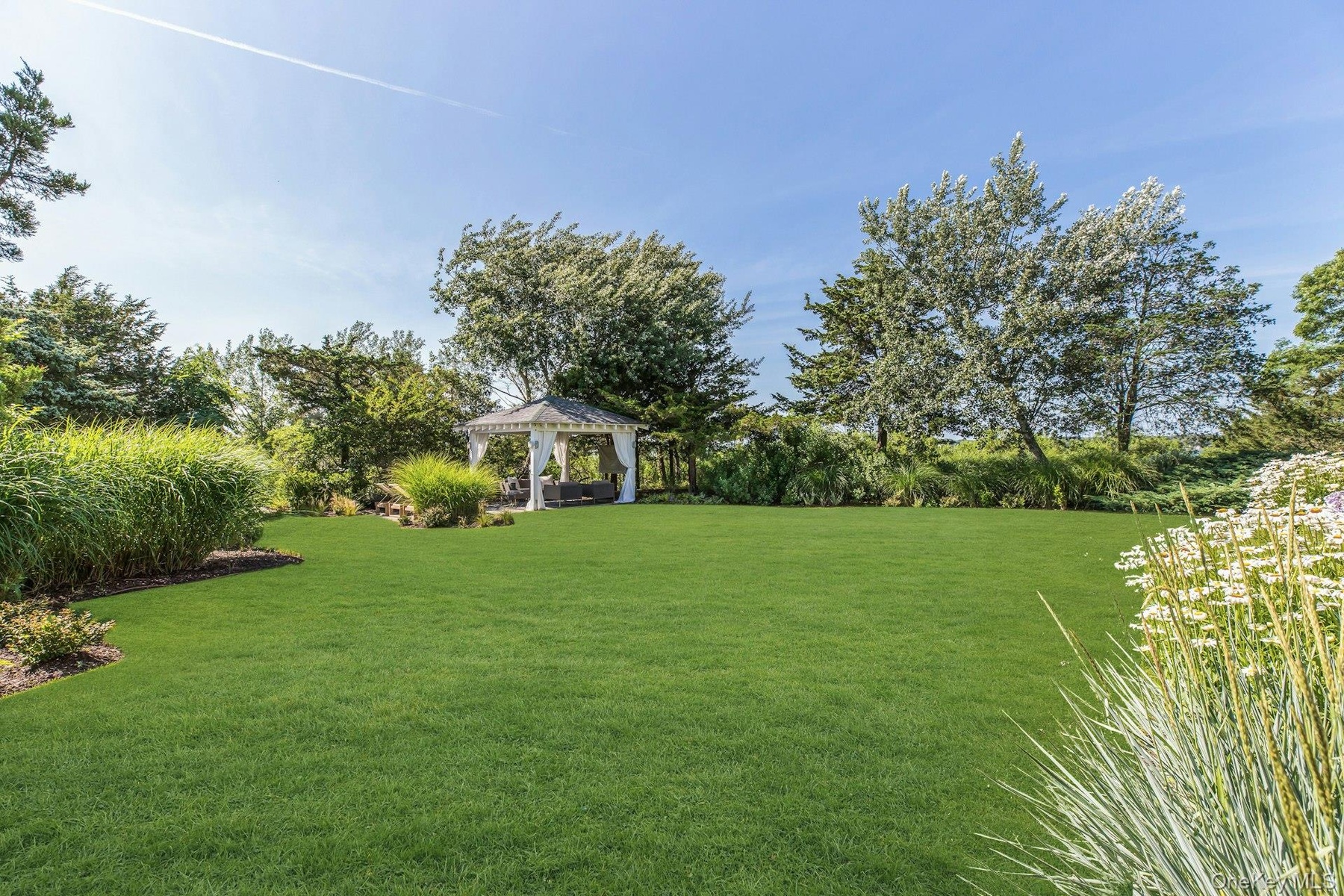 Expansive manicured lawn featuring a white gazebo with drapes, surrounded by mature trees and varied landscaping
