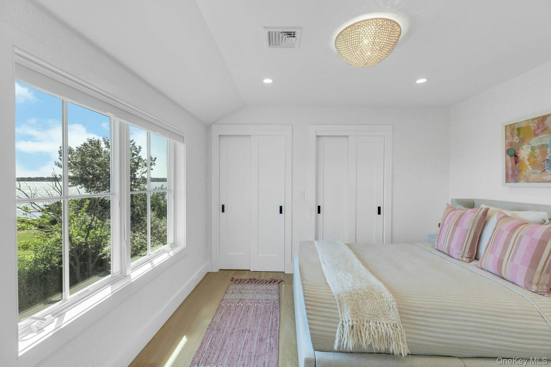 Light-filled bedroom featuring a multi-pane window overlooking a body of water and lush greenery, wood-finish flooring, two sliding closet doors with 