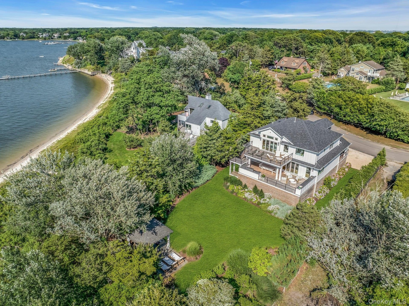 Modern coastal home with multiple outdoor decks, a manicured lawn, and mature trees, featuring a private sandy beach and a long dock extending into th