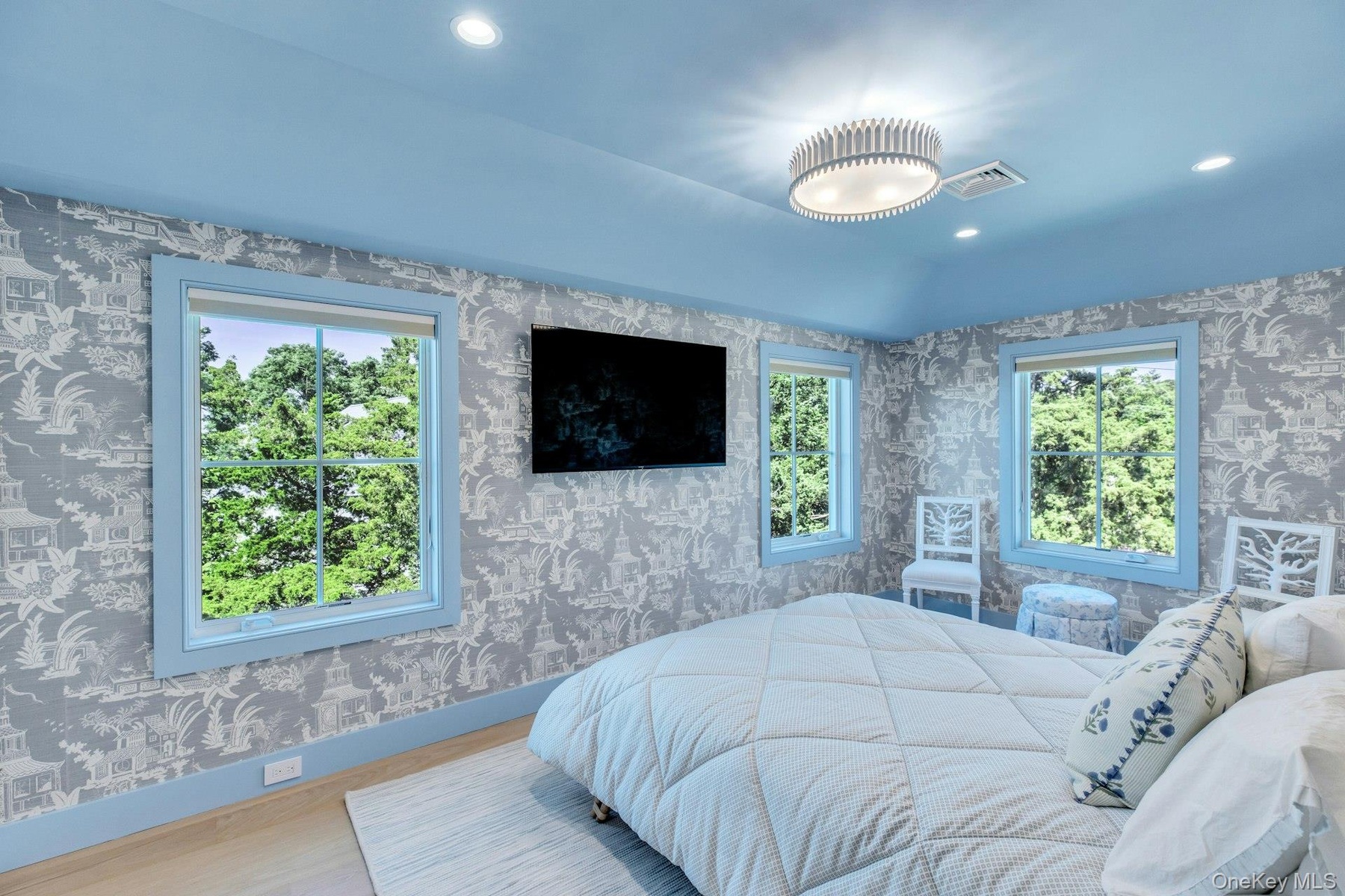 Spacious bedroom featuring light wood-finish flooring, patterned wallpaper, a built-in wall-mounted television, and multiple windows with light blue t
