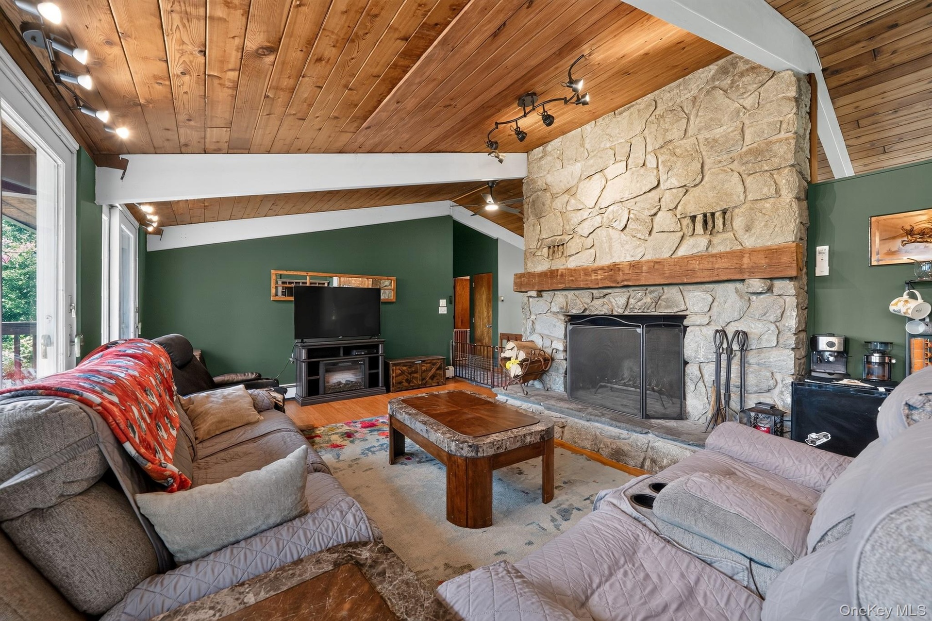 Living area featuring a stone fireplace with a substantial timber mantel, vaulted wood-paneled ceilings, hardwood-style flooring, track lighting, and 