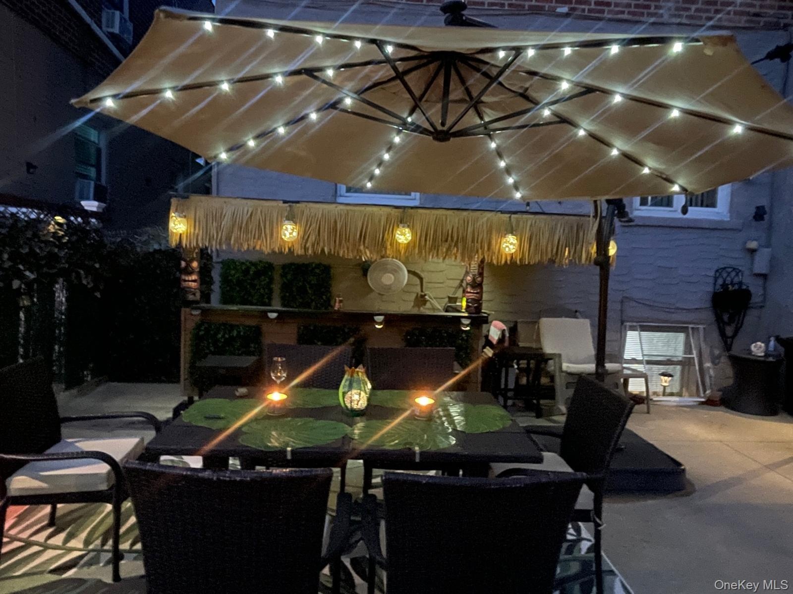 Paved outdoor patio featuring an illuminated umbrella, a built-in bar with a thatched roof accent, and a privacy screen of artificial greenery