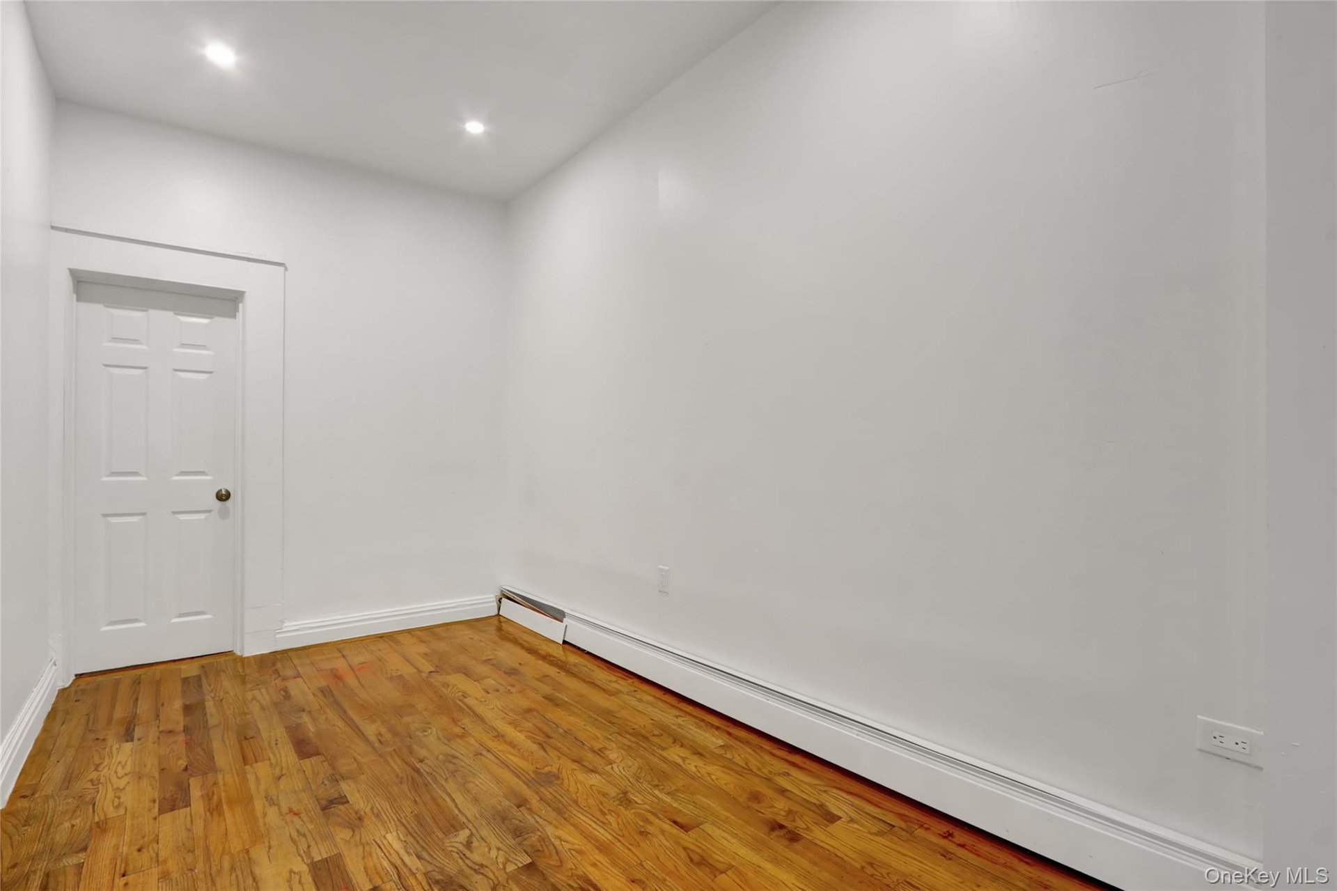 Room featuring wood-finish flooring, white walls, recessed lighting, and a white paneled door with a brass-toned knob