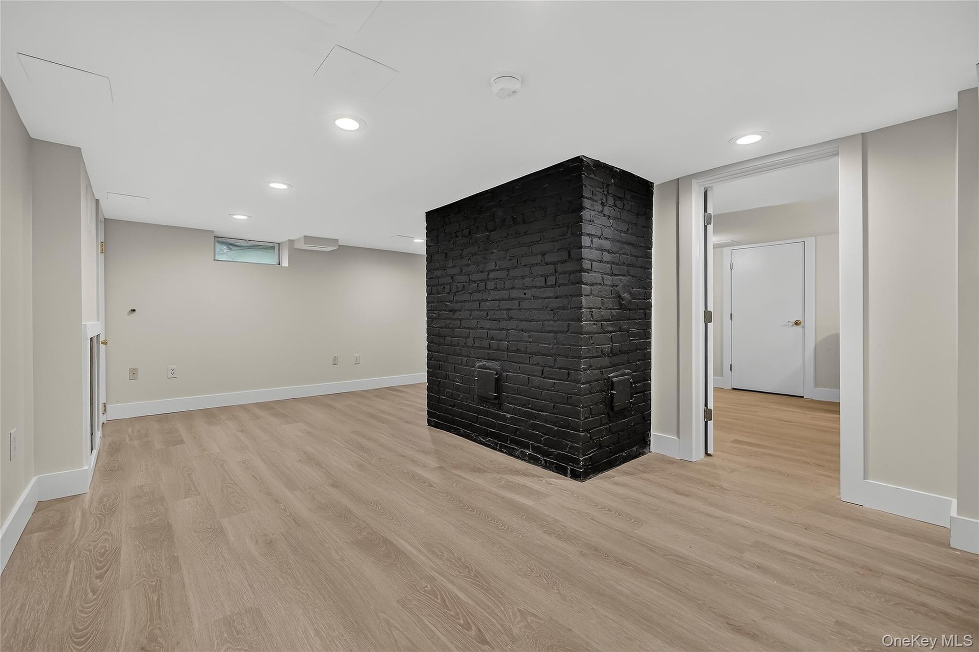 Spacious room with light wood-finish flooring, a painted brick feature, recessed lighting, and a doorway to an additional room with wood-finish floori