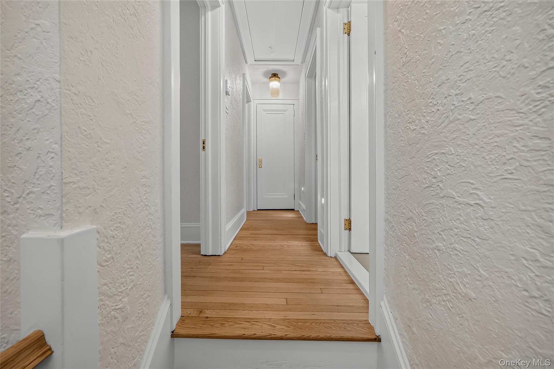 Hallway featuring wood-finish flooring, white doors with brass hardware, textured wall finishes, white trim, and a ceiling-mounted light fixture