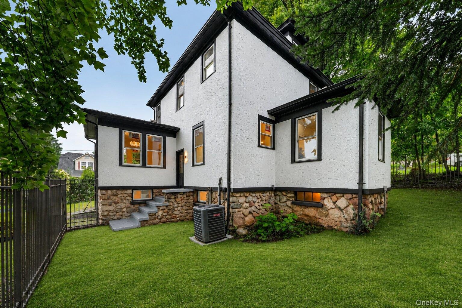 Rear exterior showcasing white stucco siding with black trim, a stone foundation, a manicured lawn, and a black wrought iron fence