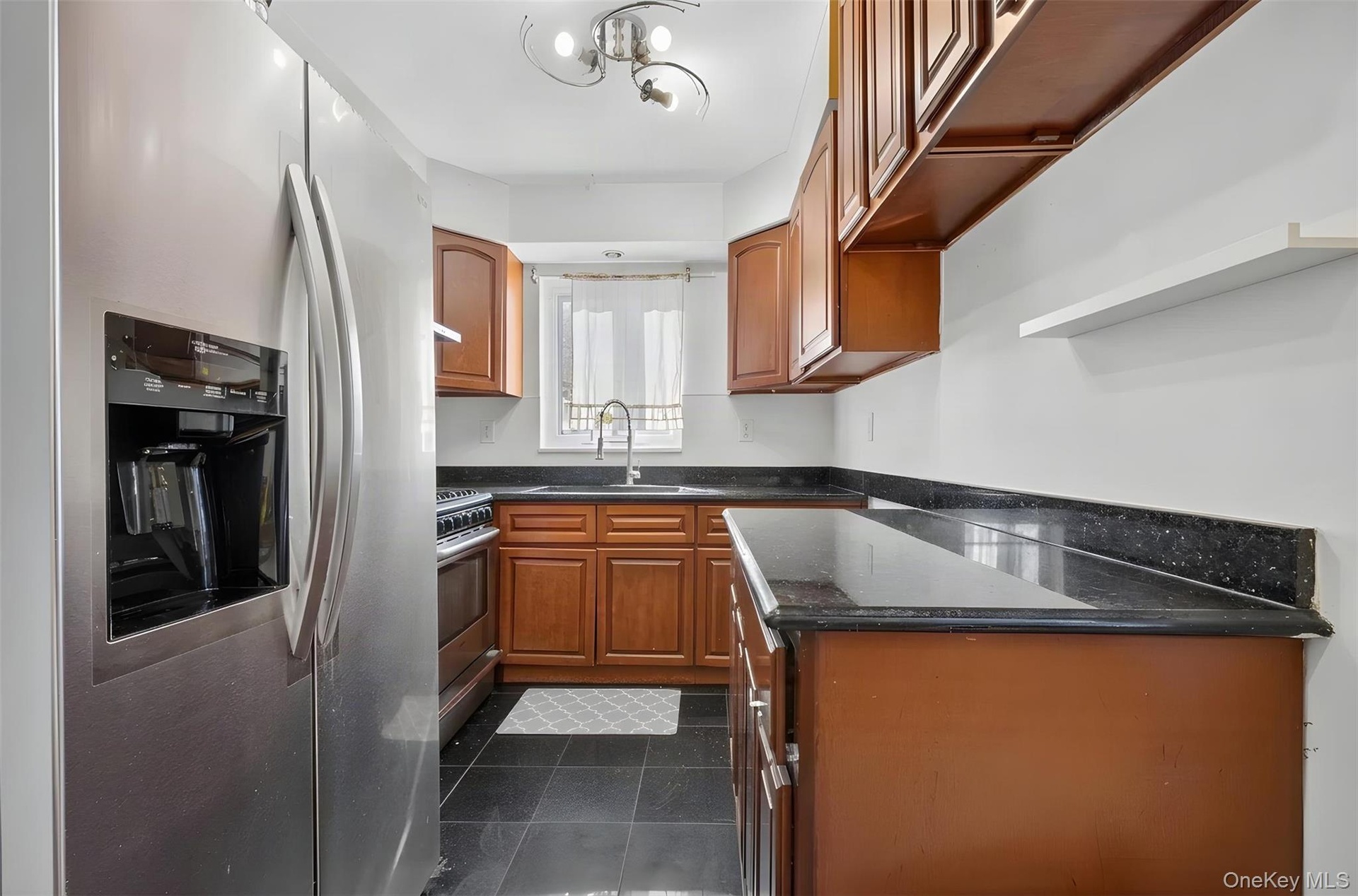 Stainless steel refrigerator with a dispenser, black tile flooring, wood-finish cabinetry, dark speckled countertops, and a stainless steel sink with 