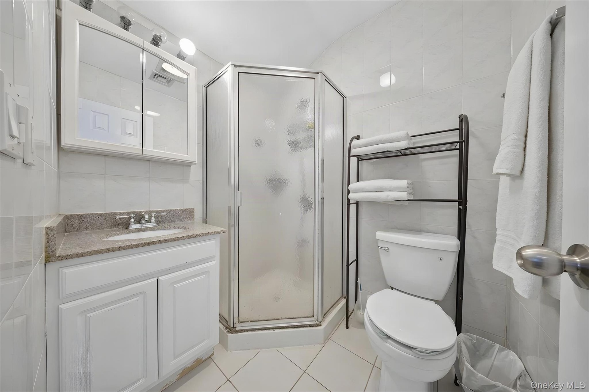 White tiled bathroom featuring a corner shower, white vanity with a light-toned countertop, and a mirrored medicine cabinet