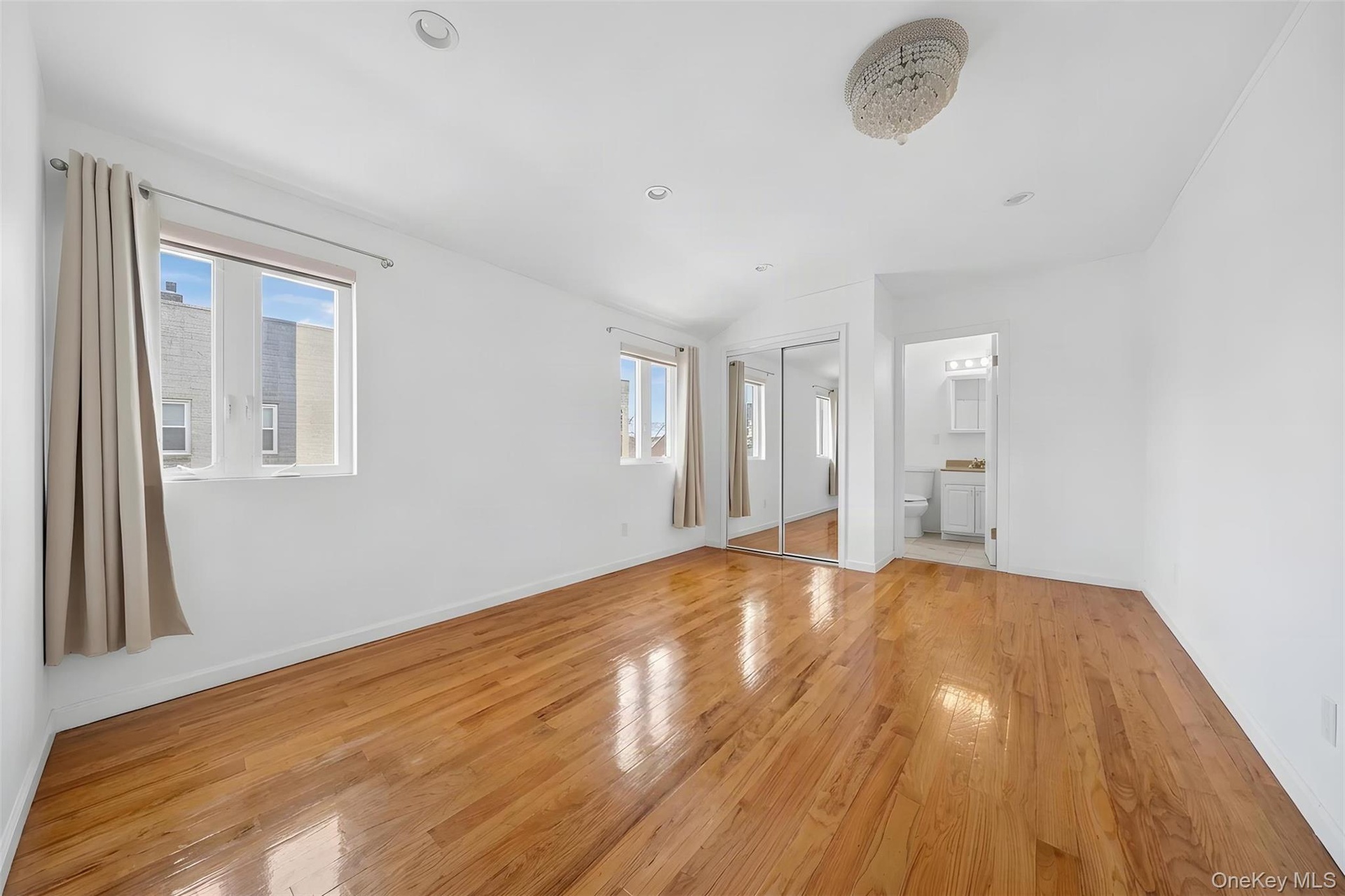 Bright and spacious room featuring gleaming wood-finish flooring throughout, abundant natural light from multiple windows with soft drapery, crisp whi