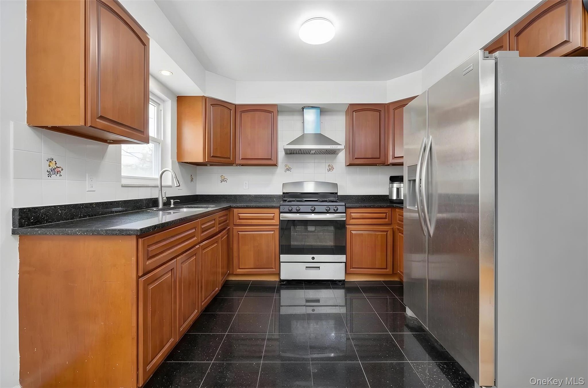 Kitchen featuring wood cabinetry, dark countertops, a stainless steel refrigerator, a range, and a stainless steel range hood