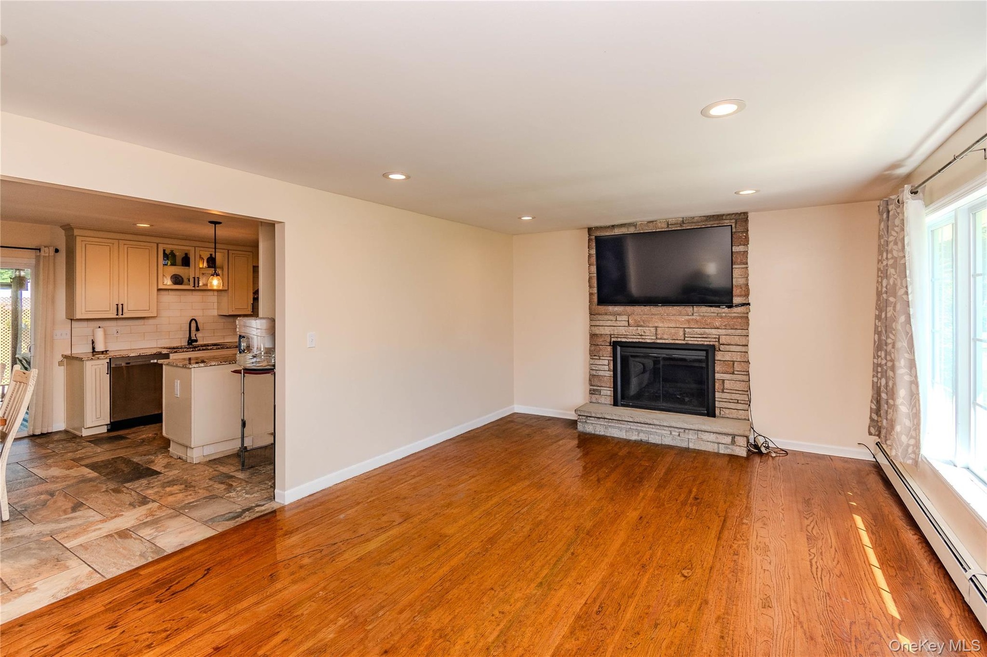 Hardwood flooring, recessed lighting, stone-clad fireplace, large window, and an open pass-through to the kitchen