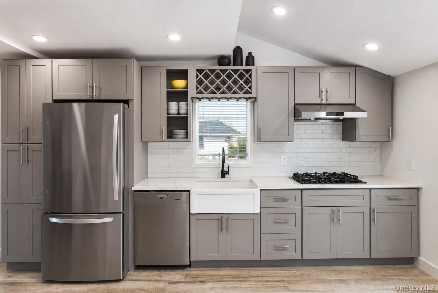 Stainless steel appliances, white subway tile backsplash, white farmhouse sink, grey shaker-style cabinetry, and wood-finish flooring