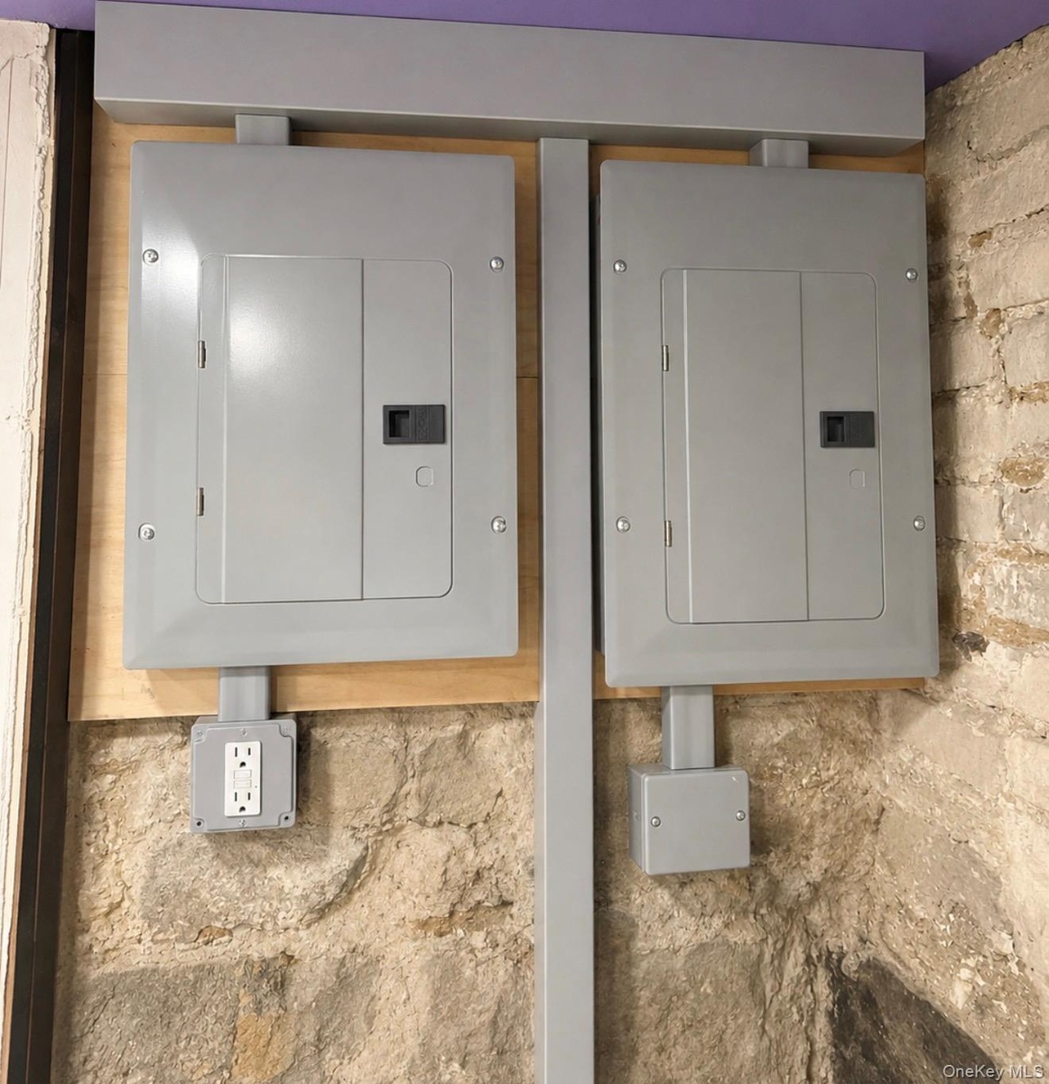 Electrical panels with a wall outlet, mounted on a wood-finish backboard, set against a natural stone wall