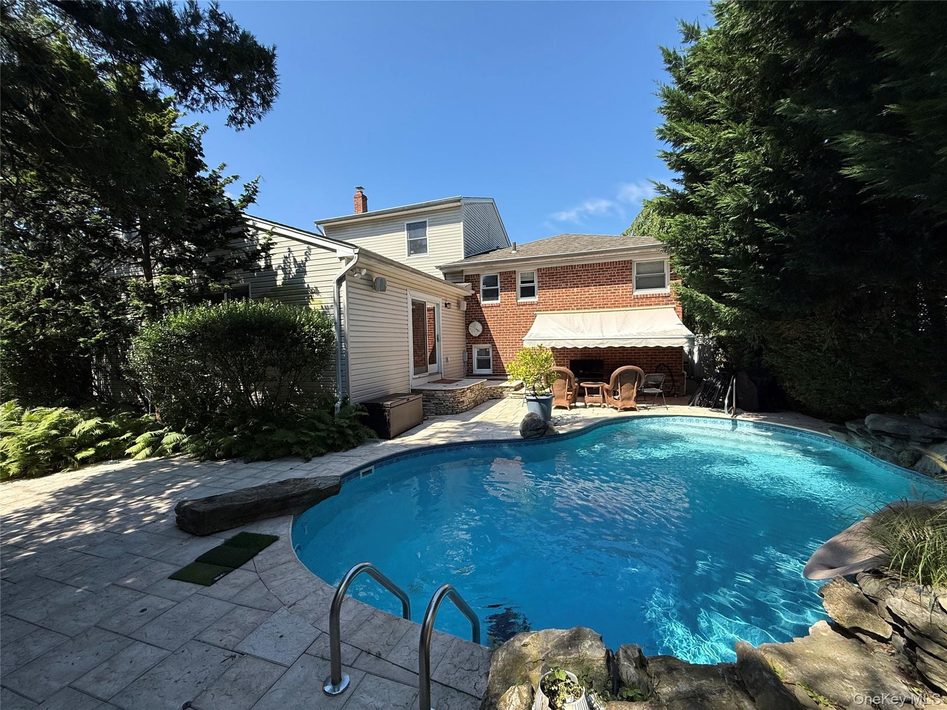 Backyard oasis featuring a free-form in-ground pool surrounded by a stone patio. Retractable awning shades a cozy seating area, perfect for entertaini
