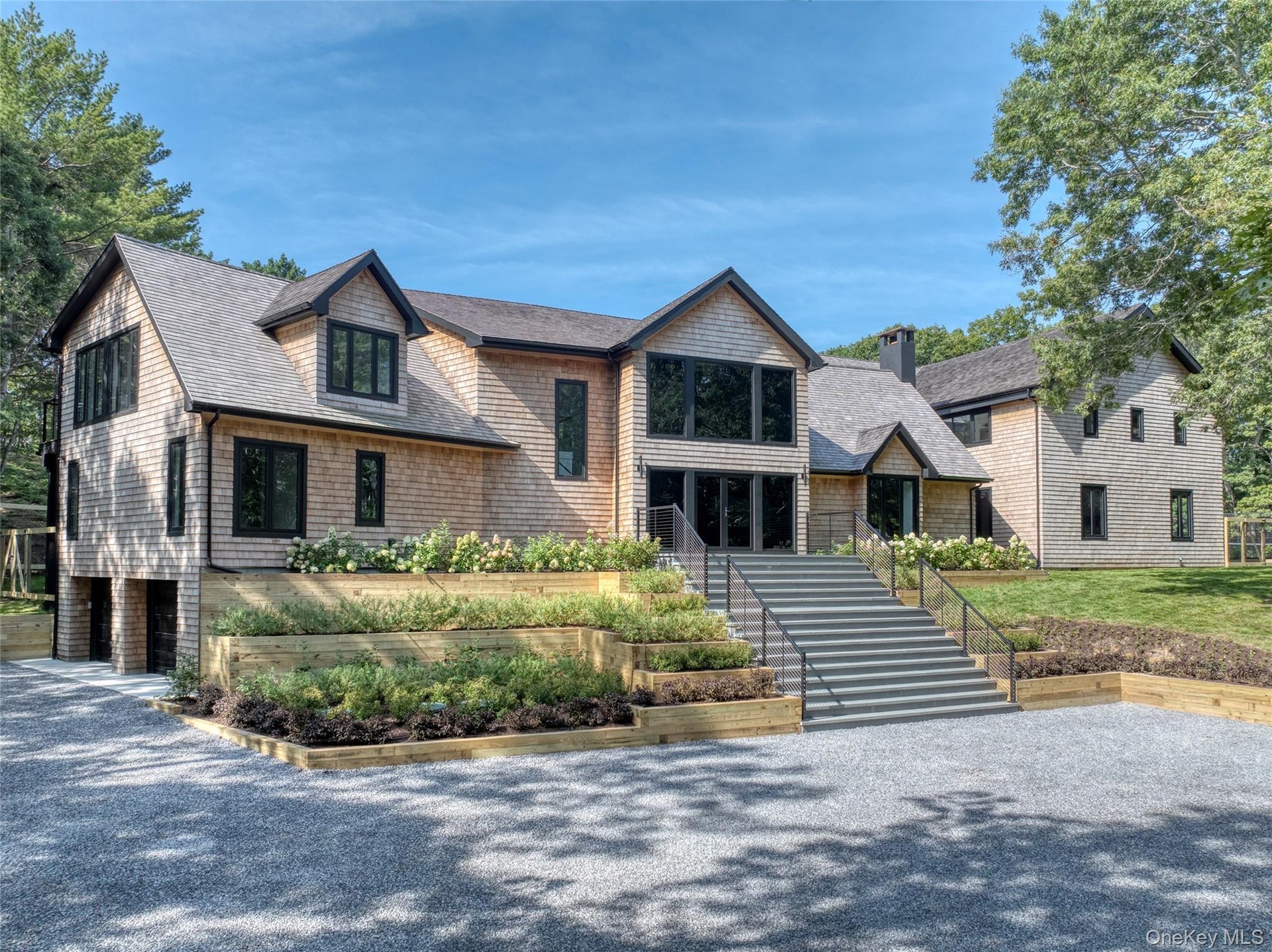 Shingle-style exterior with black trim windows, multi-level tiered landscaping, broad front steps, and a gravel driveway