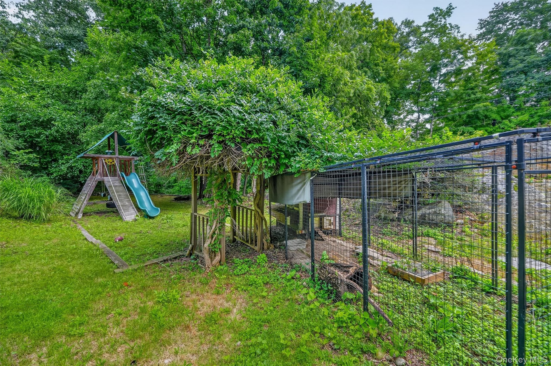 Lush backyard featuring a wooden playset with a slide, a gazebo structure with natural wood posts, and a fenced enclosure ( Chicken Coup)