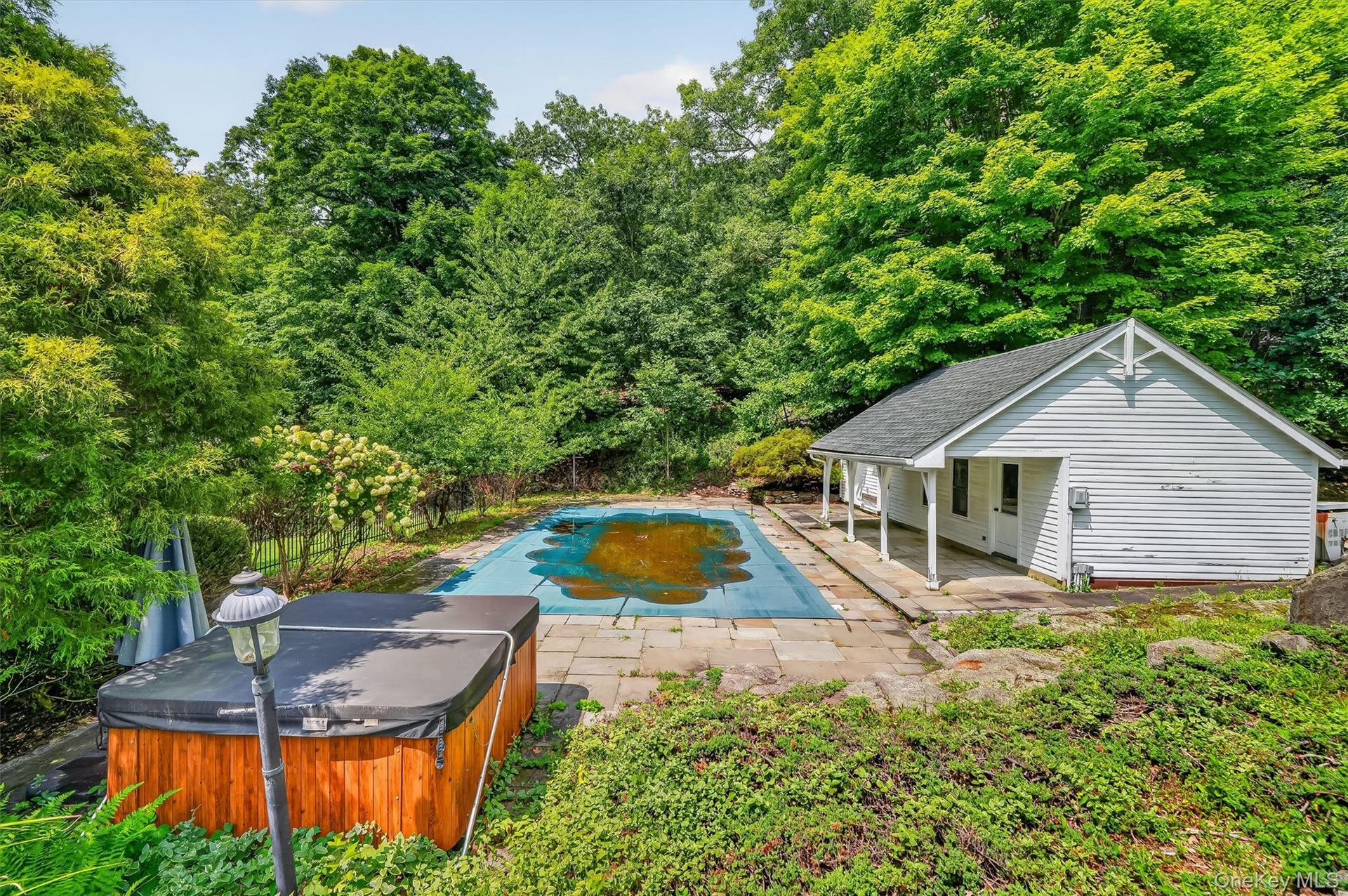 Outdoor swimming pool with surrounding stone patio, white siding pool house with covered porch, and wood-sided hot tub
