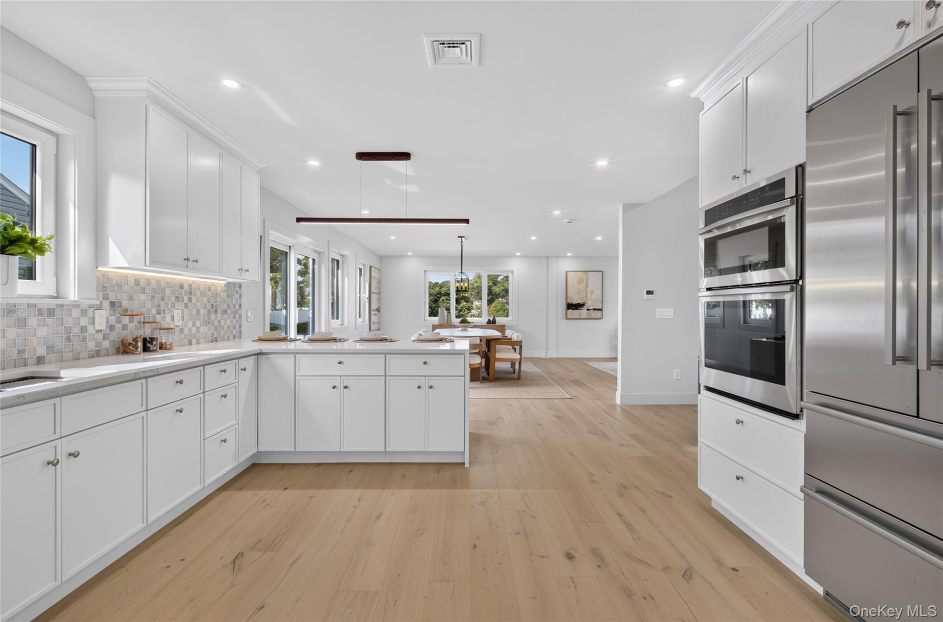 Open-concept kitchen featuring extensive white cabinetry, light wood-finish flooring, stainless steel appliances, and a mosaic tile backsplash