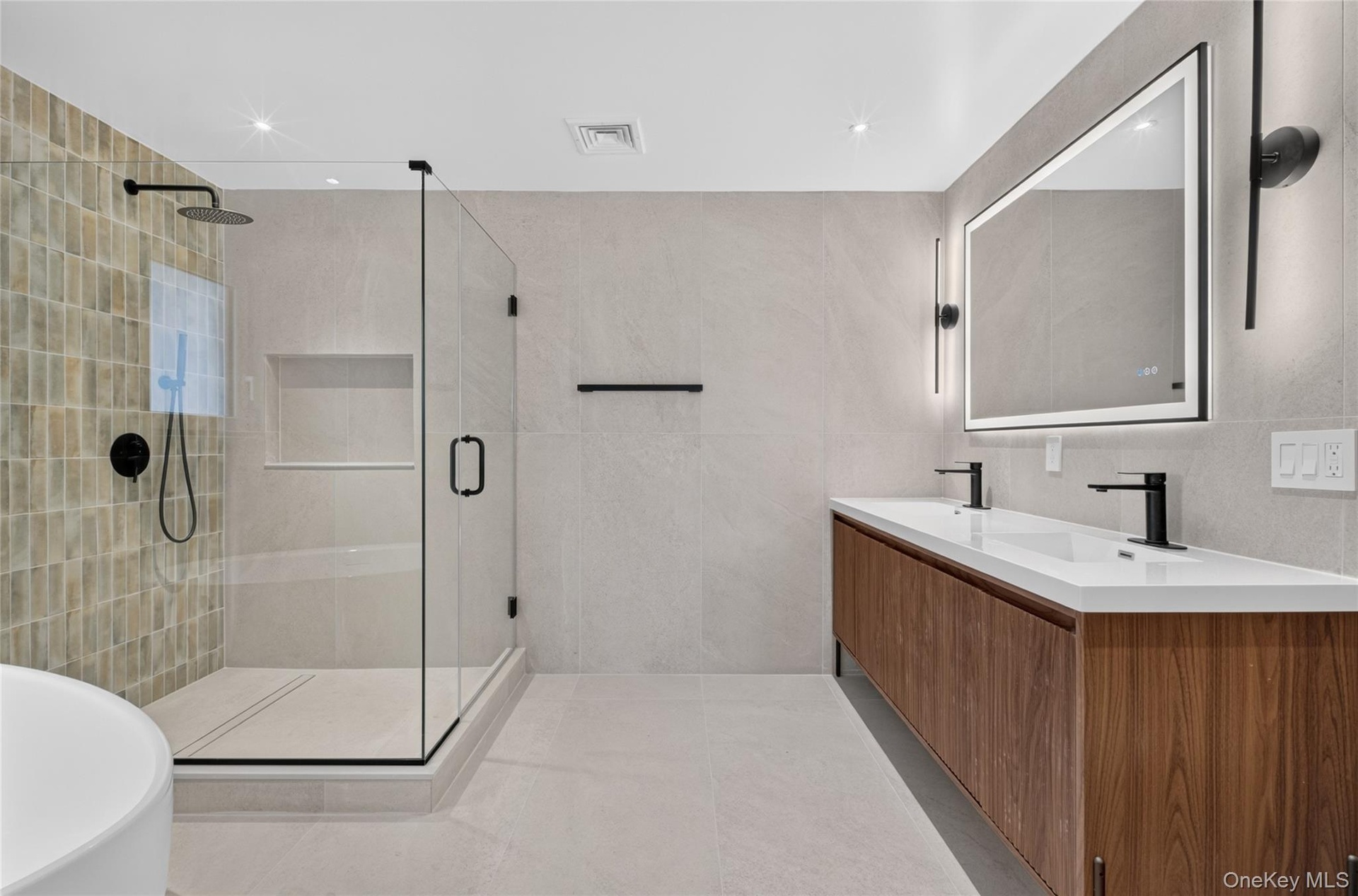 Glass-enclosed shower with a tiled wall and integrated niche, dual vanity with wood-finish cabinetry, and an illuminated mirror with modern sconces