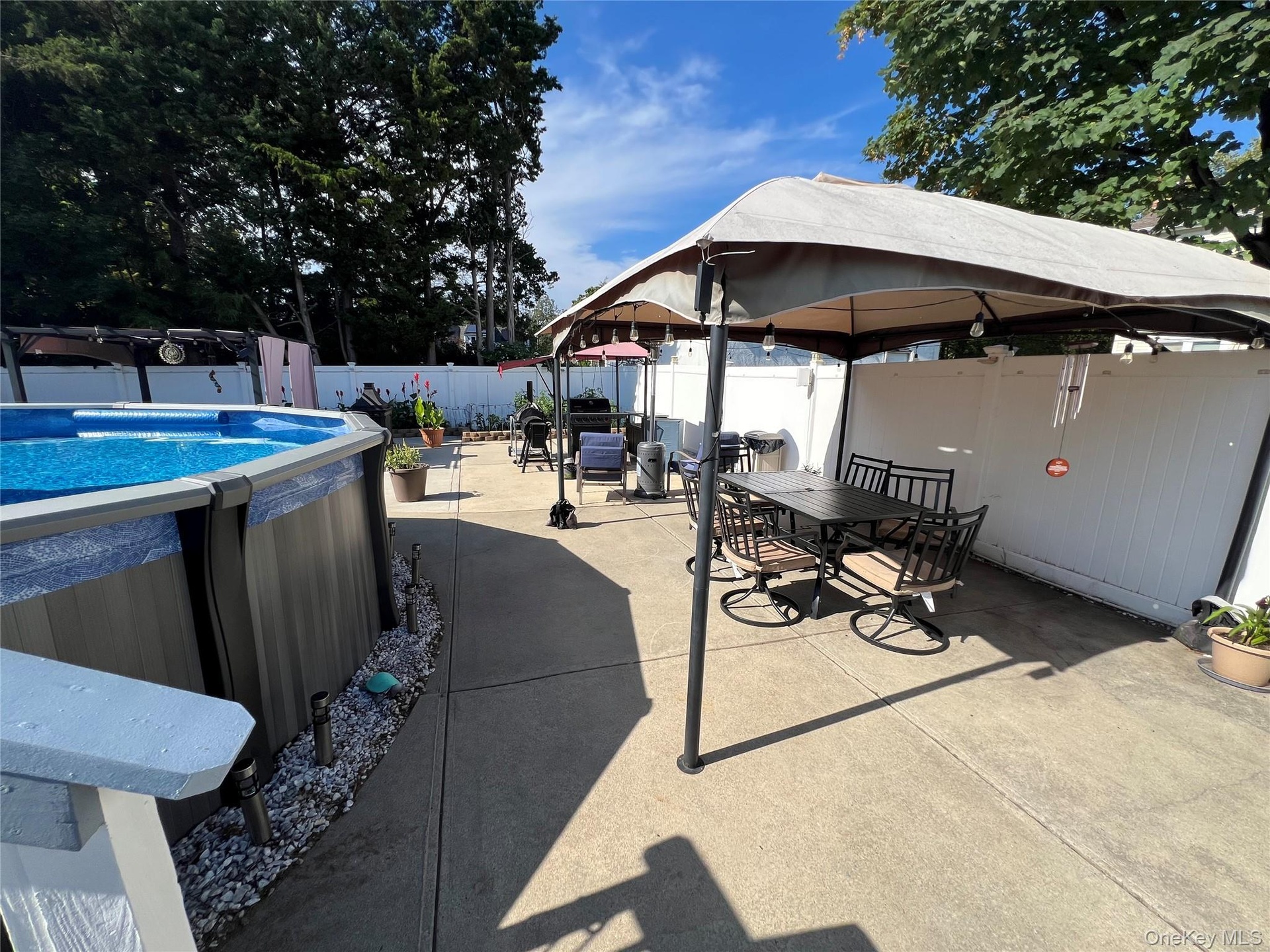 Expansive concrete patio with an above-ground pool, white privacy fencing, and mature perimeter trees