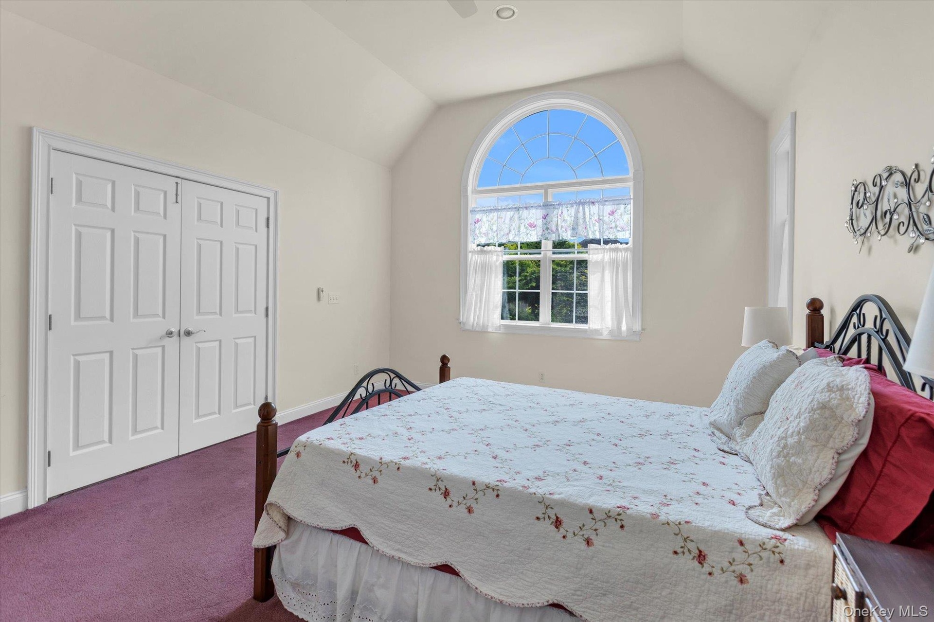 Bright bedroom suite featuring an arched transom window with plantation shutters, vaulted ceiling, wood-finish closet doors
