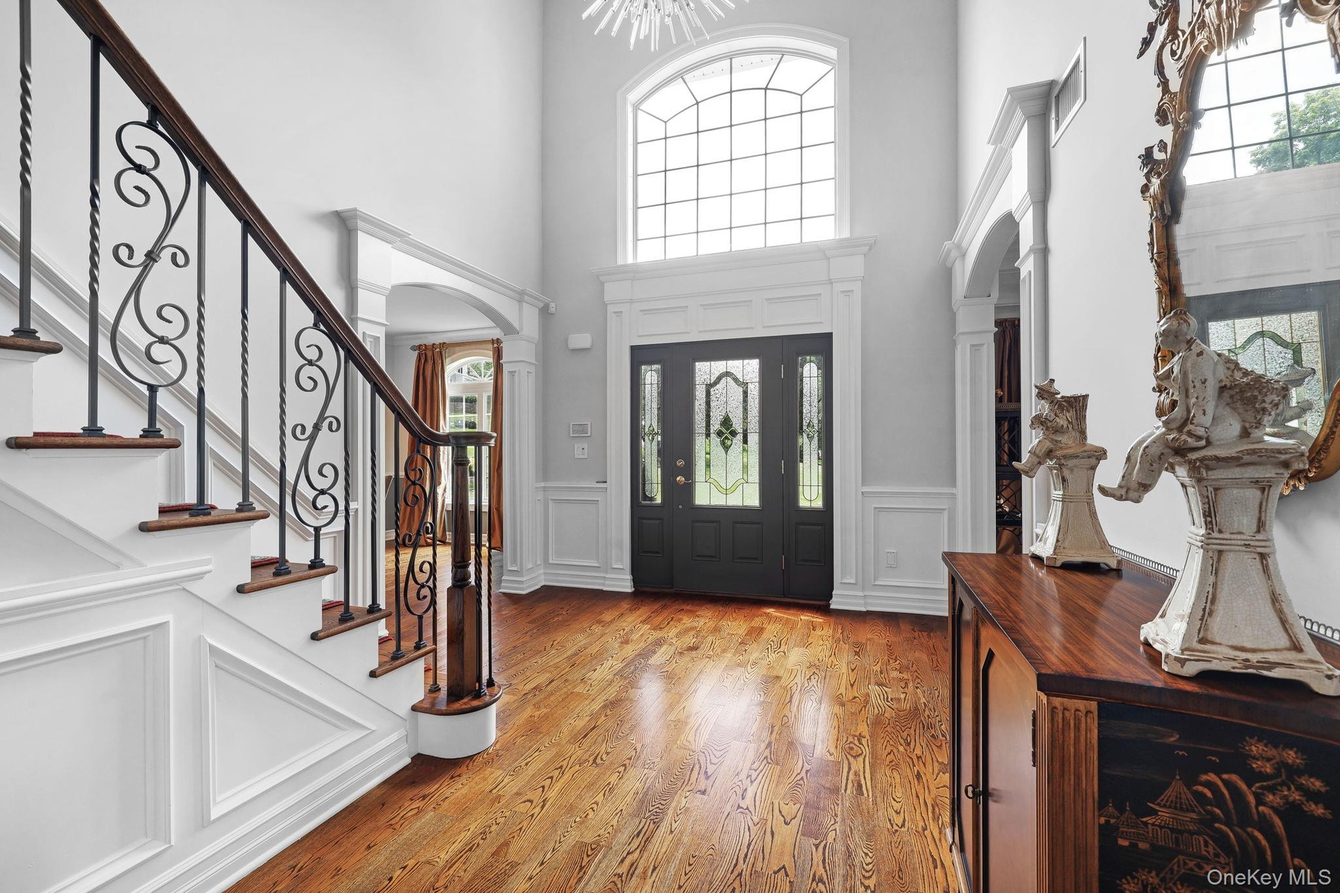 Two-story entryway with a decorative arched window, dark wood-finish flooring, and a staircase featuring intricate wrought iron balusters