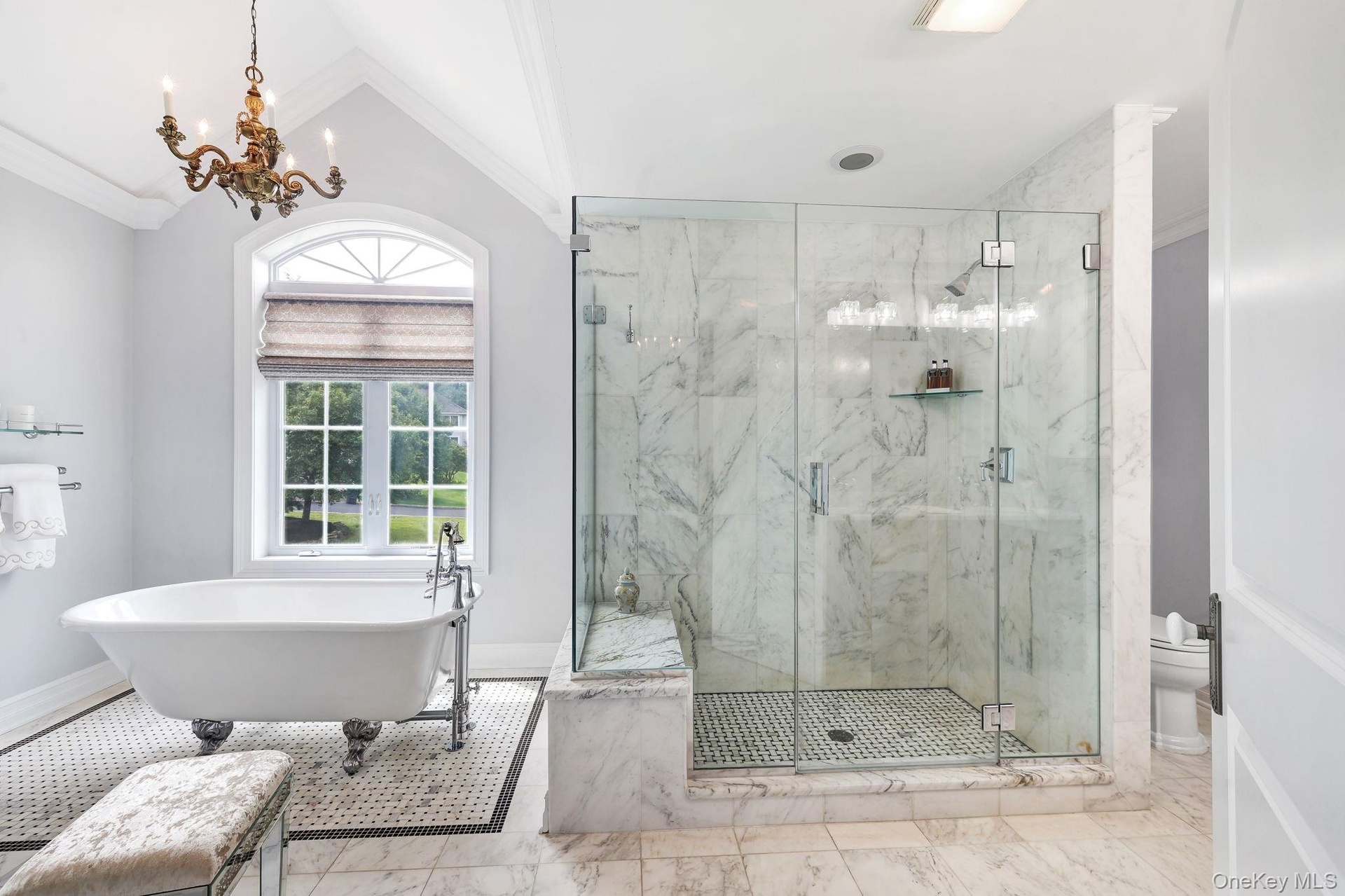Clawfoot soaking tub, glass-enclosed shower with marble-look tile, arched window, chandelier, and a mosaic tile floor accent