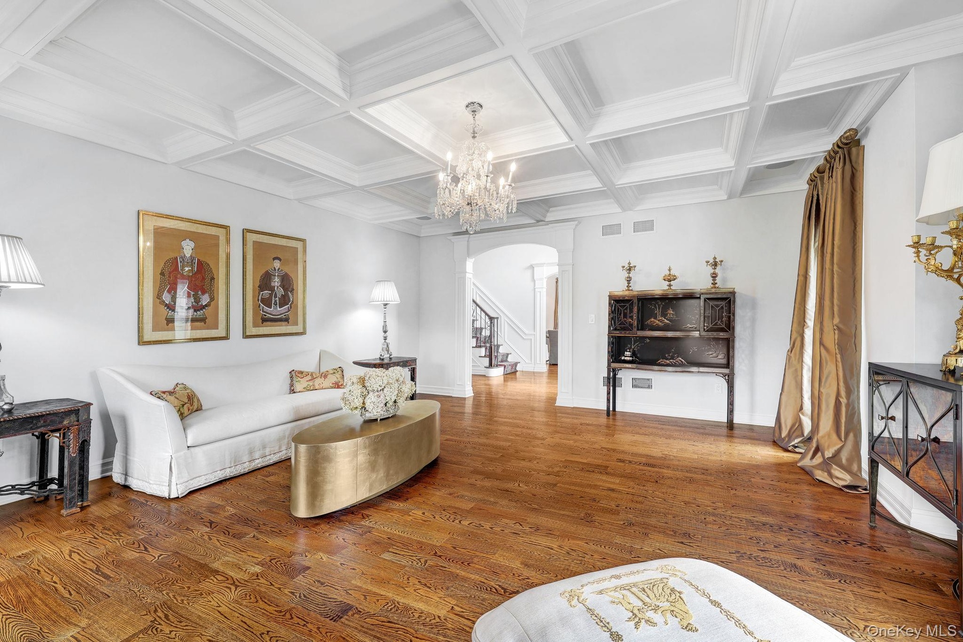 Hardwood flooring and a coffered ceiling with a crystal chandelier illuminate the living space, featuring an arched doorway and a built-in display cab