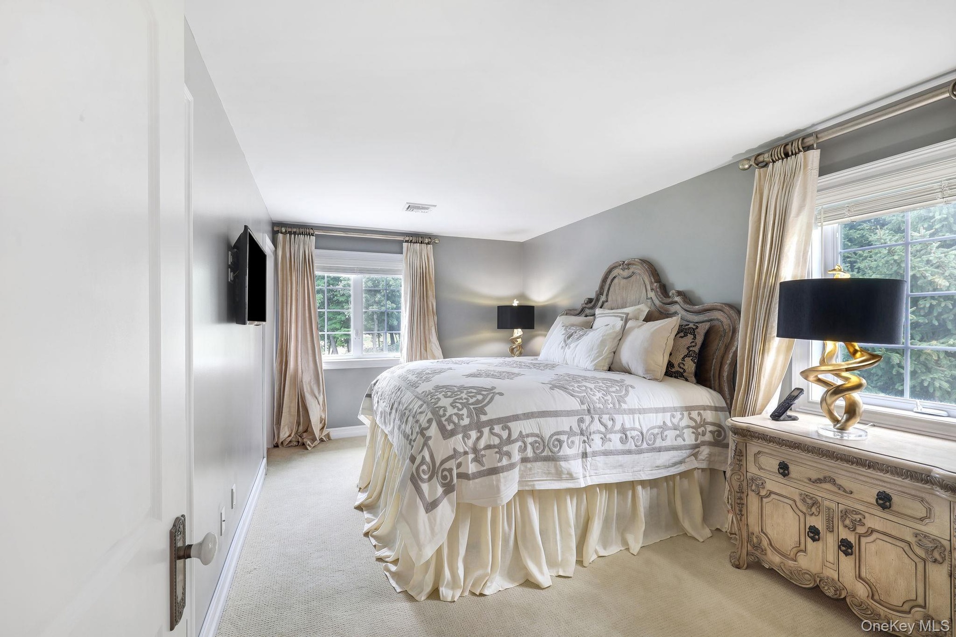 Wall-mounted television with an ornate headboard, light gray walls, and large windows