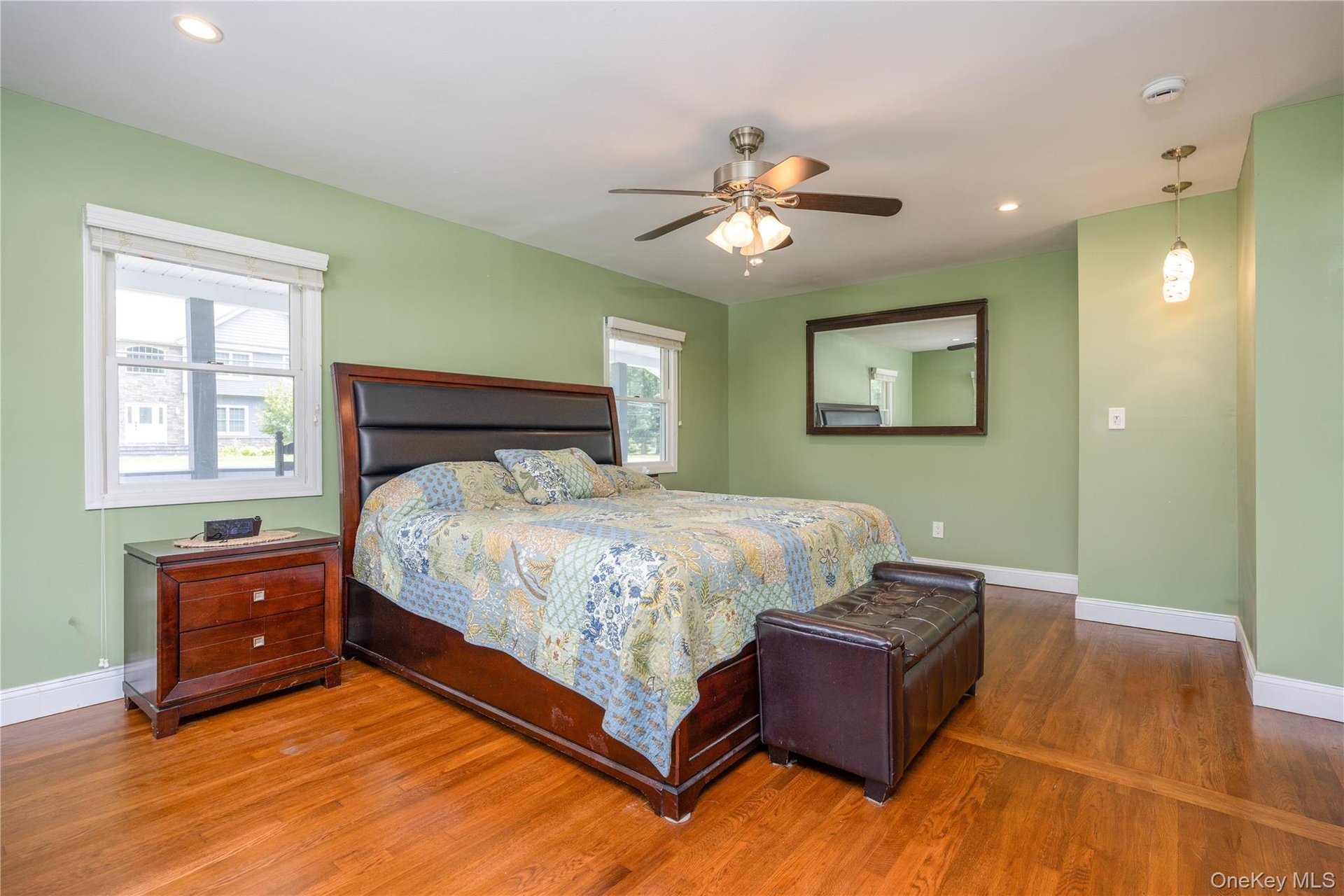 Spacious primary bedroom showcasing soft green walls, hardwood flooring, ceiling fan with integrated lighting, and a framed mirror