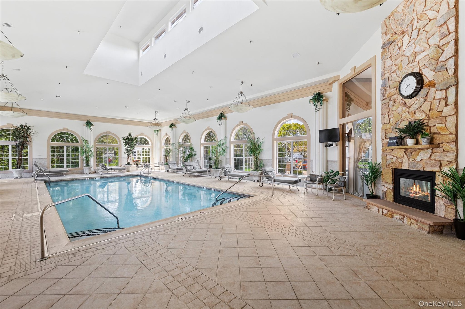 Indoor swimming pool with integrated spa, large arched windows, stone-clad fireplace with hearth, and tiled flooring