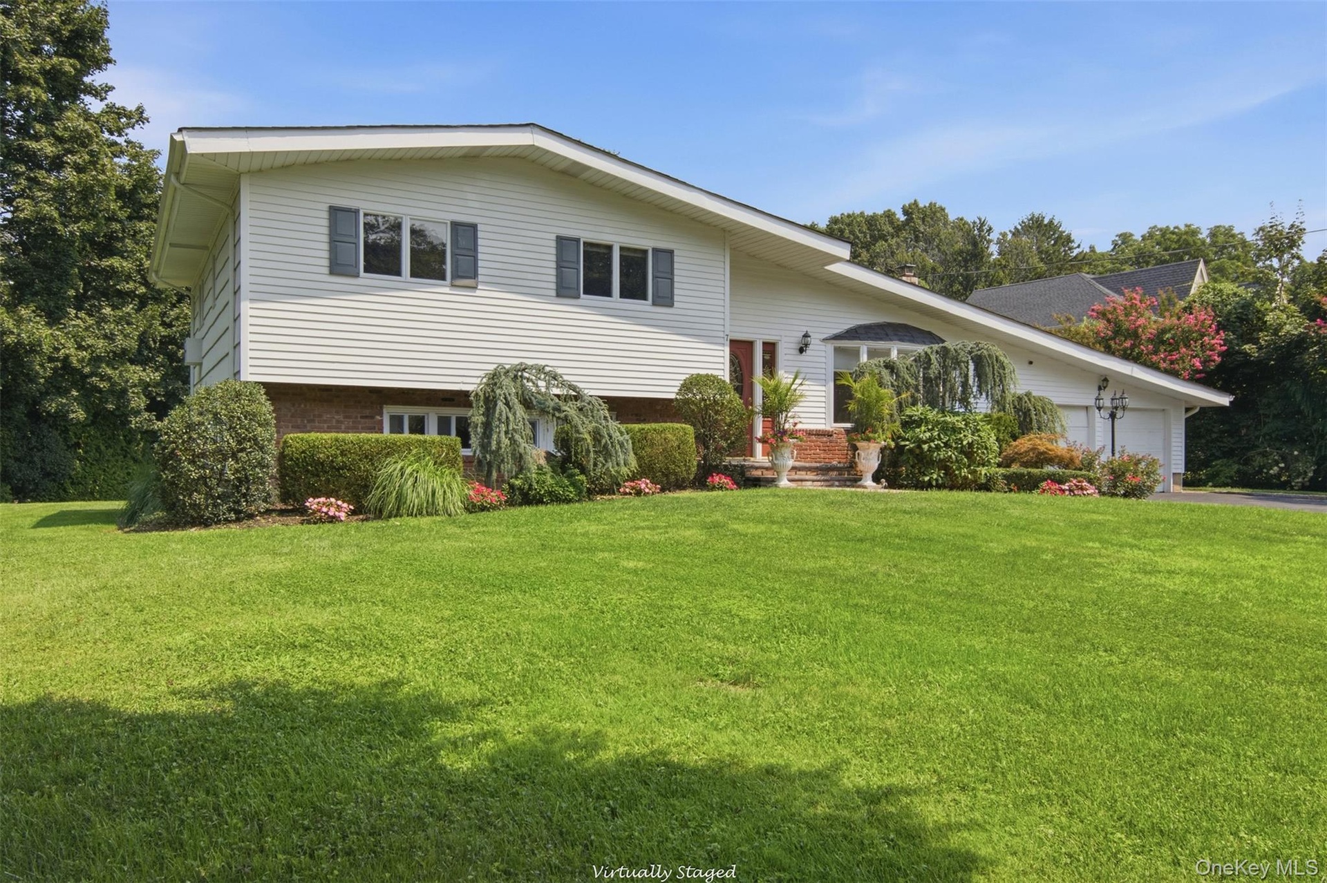 White siding exterior with a brick lower level, featuring a two-car garage, mature landscaping, and an expansive lawn