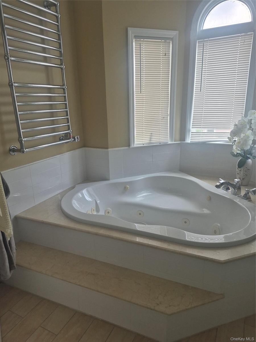 Master Bathroom with corner jetted bathtub with a light-toned surround, white tile wainscoting, an arched window and heated towel rack.