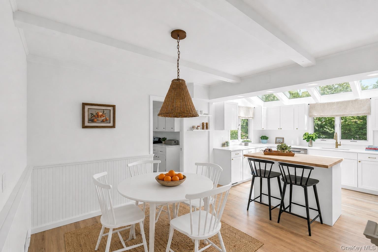 White shaker cabinetry, light wood-finish flooring, exposed ceiling beams, a central island with wood butcher block countertop, and multiple skylights